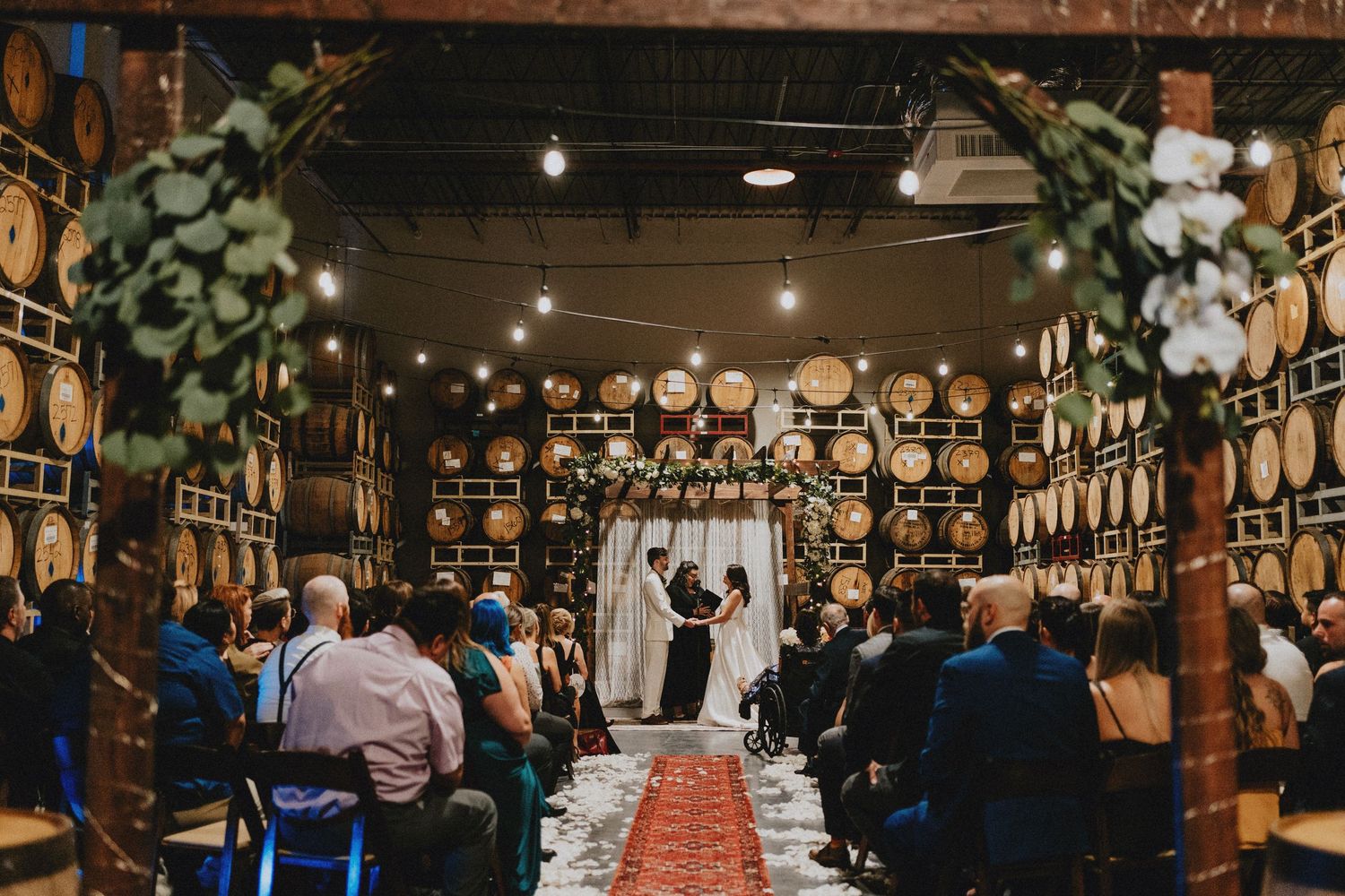 Wedding ceremony in a rustic winery with barrel walls, string lights, floral arch, and guests seated on chairs along a red aisle runner.