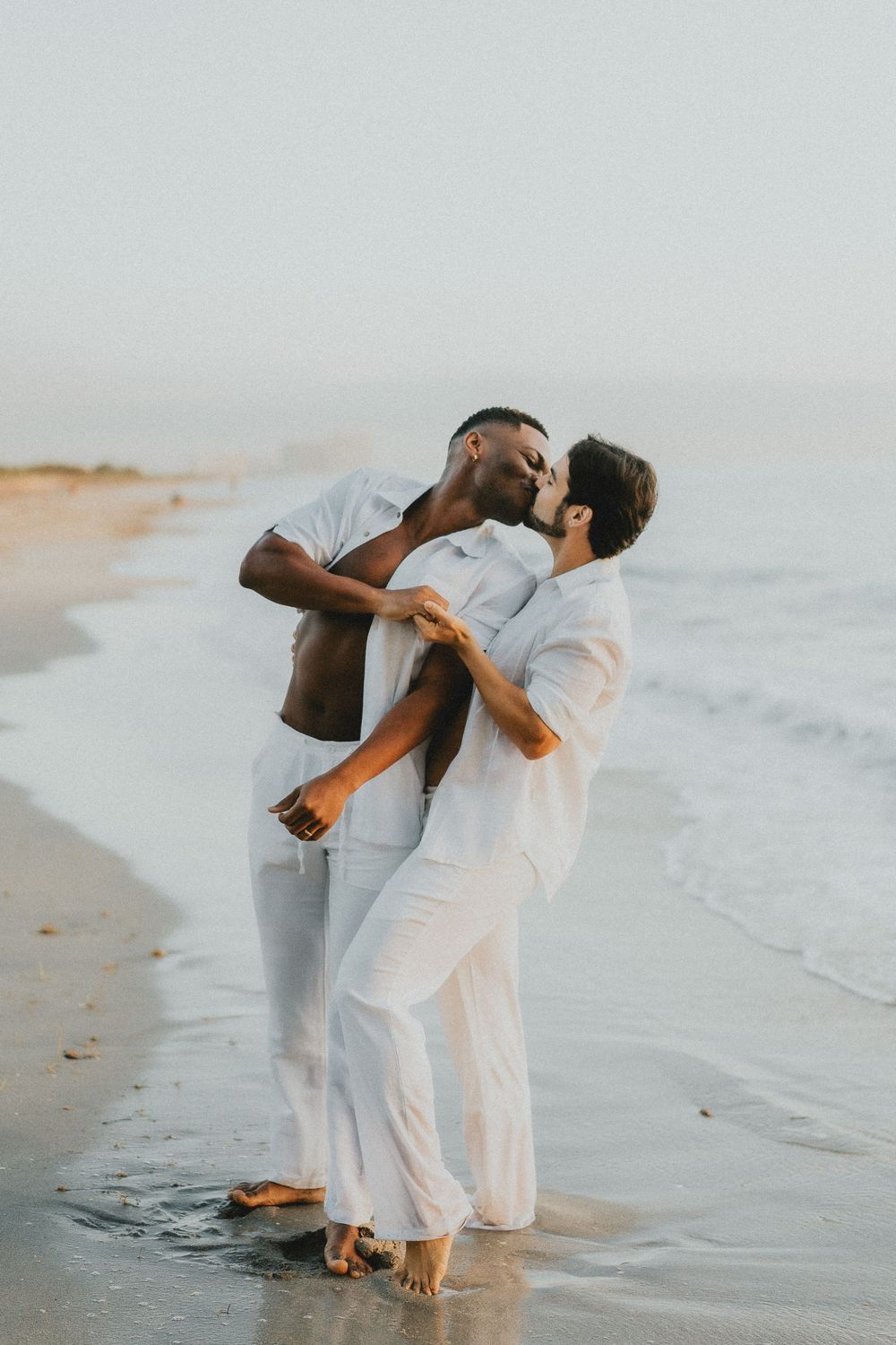 Couple in white outfits dancing playfully together on a beach at sunset.