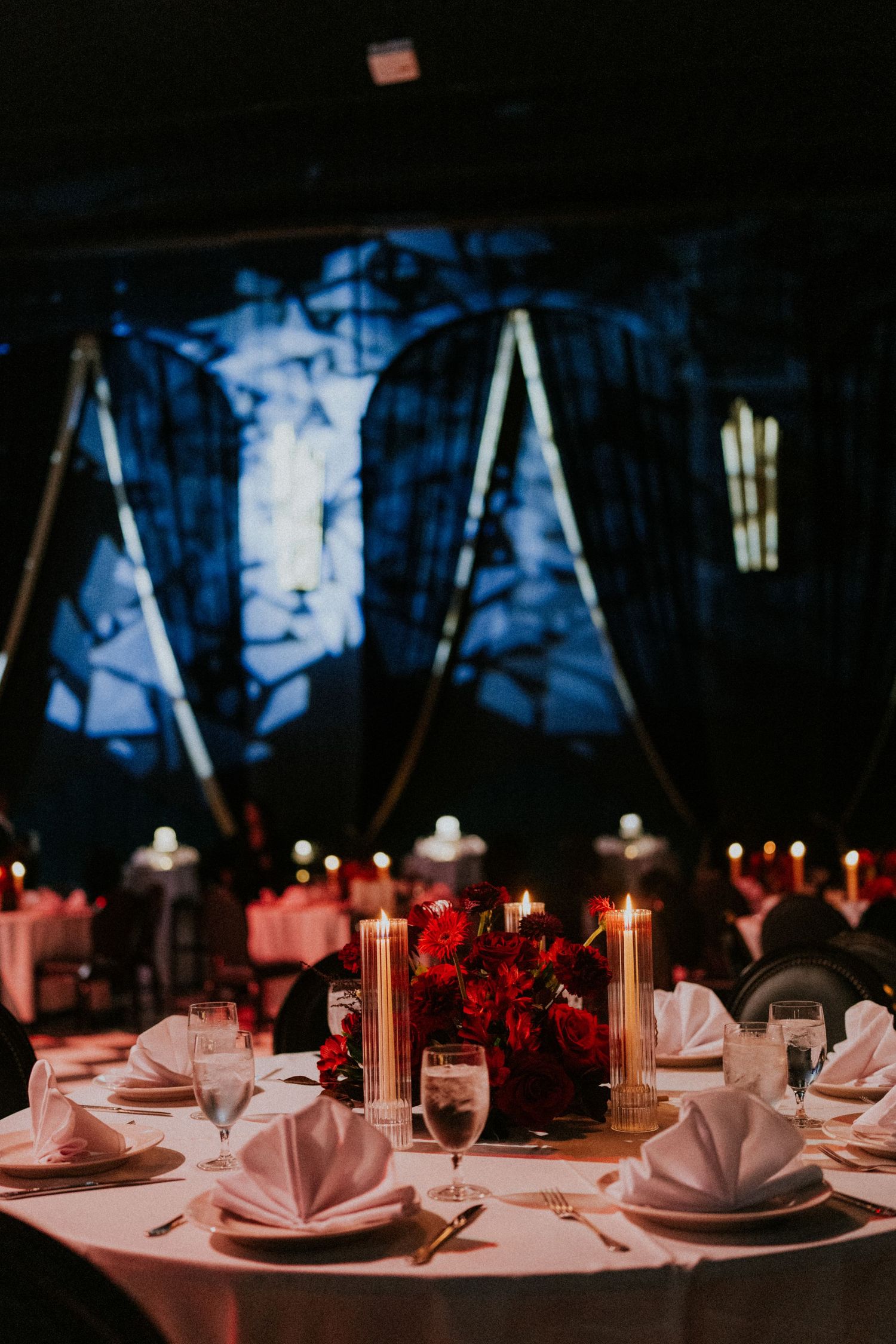 Elegant wedding reception table with white napkins, red floral centerpiece, and glowing candles in a dimly lit venue.