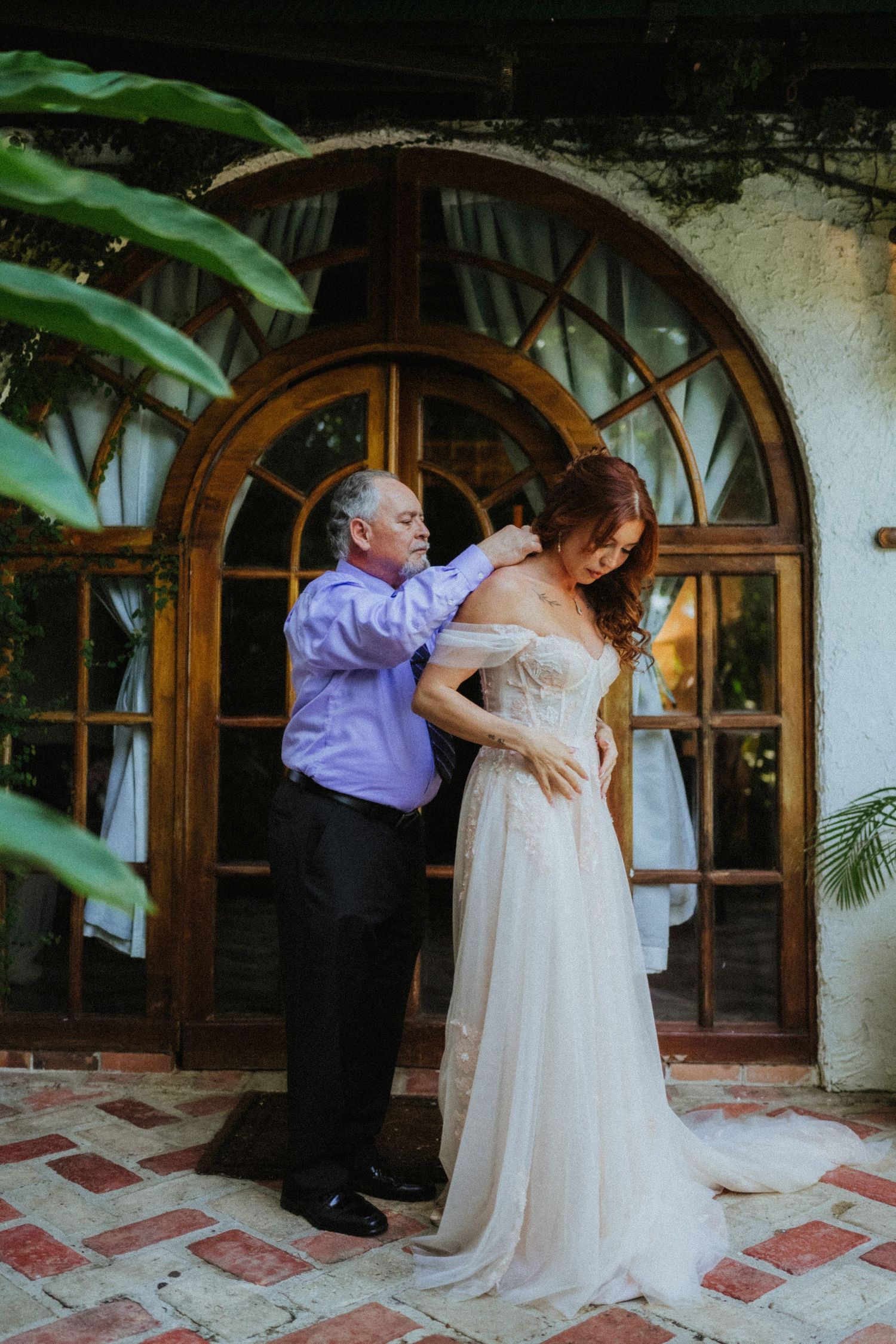 Father helping bride with wedding dress outside arched wooden doors at elegant venue.