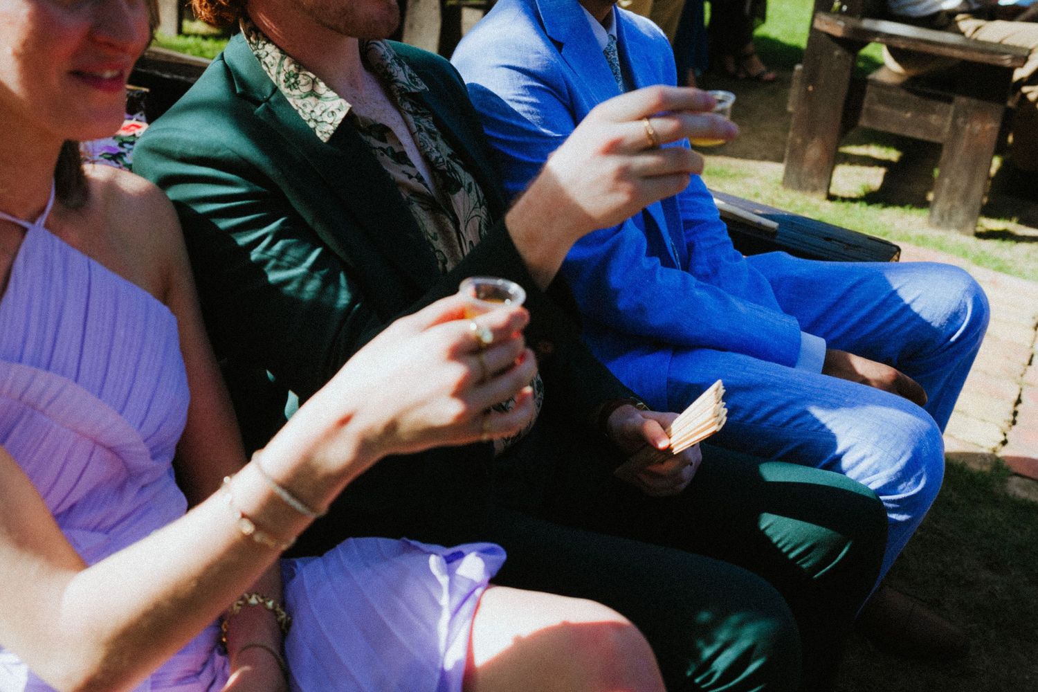 Formally dressed guests in purple and blue outfits holding drinks at an outdoor wedding reception.