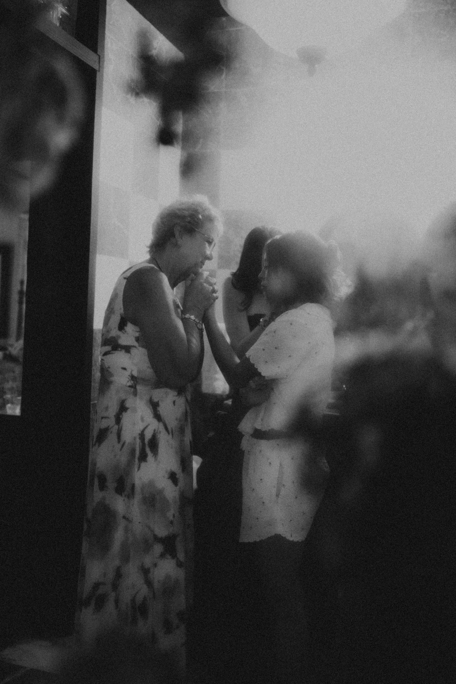 Black and white photo of women in floral dresses conversing at an indoor social gathering or event.