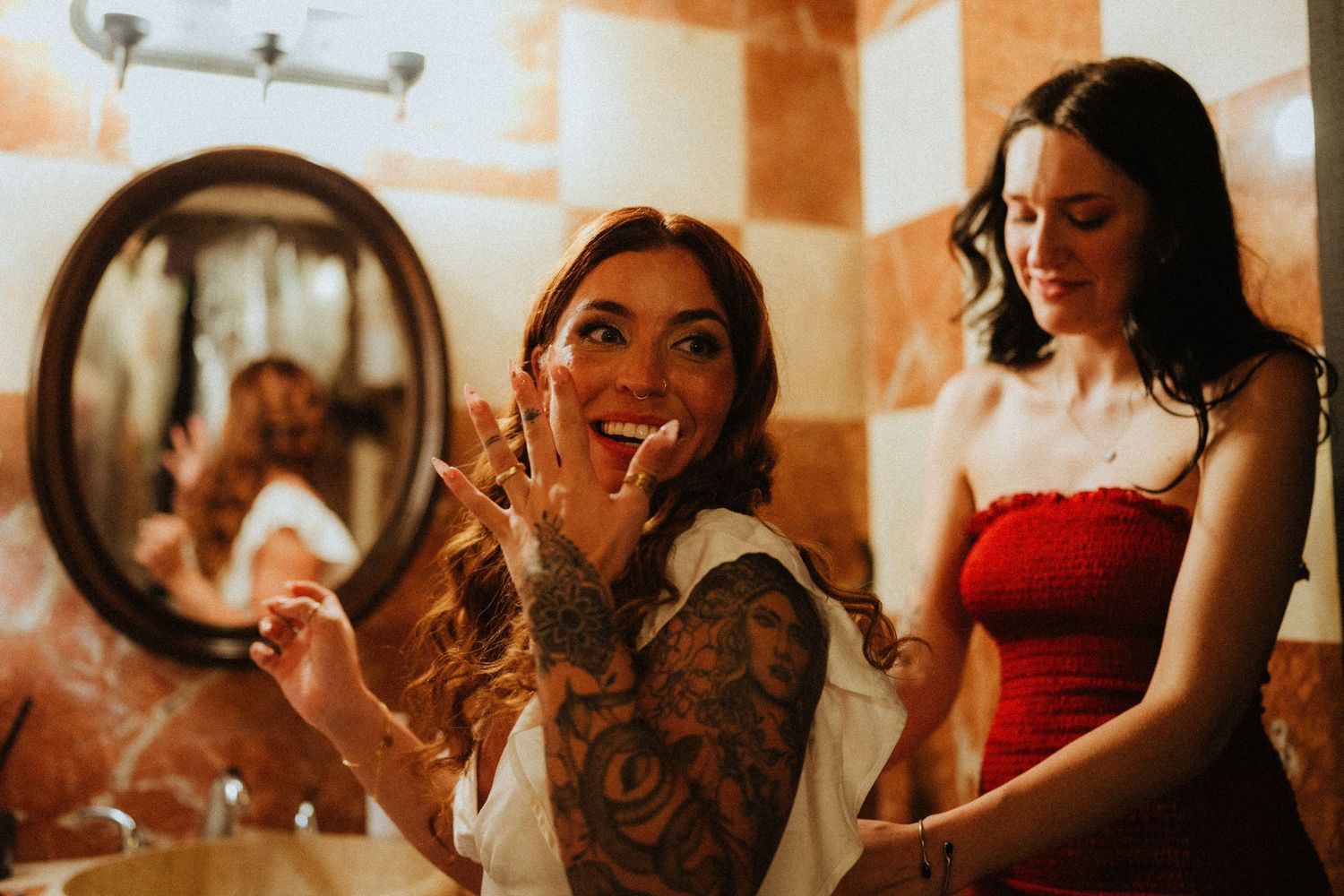 Tattooed bride applies lipstick in bathroom mirror while bridesmaid in red dress smiles nearby.