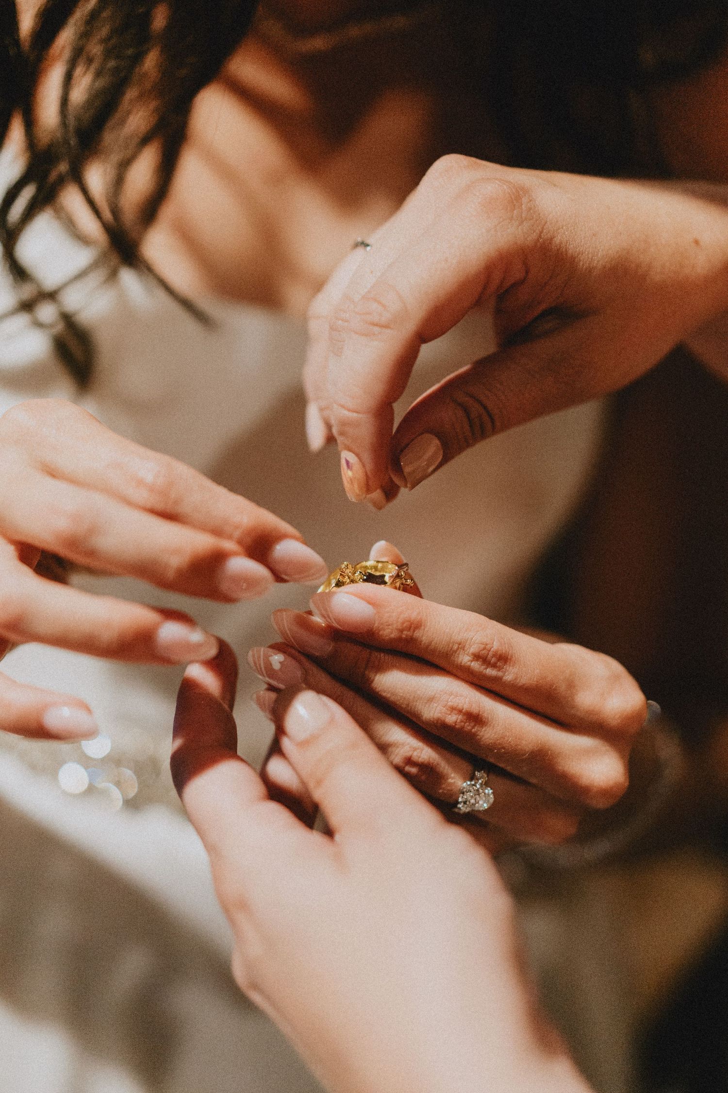 Two women examining a gold ring, one wearing a diamond engagement ring, close-up of hands with manicured nails.