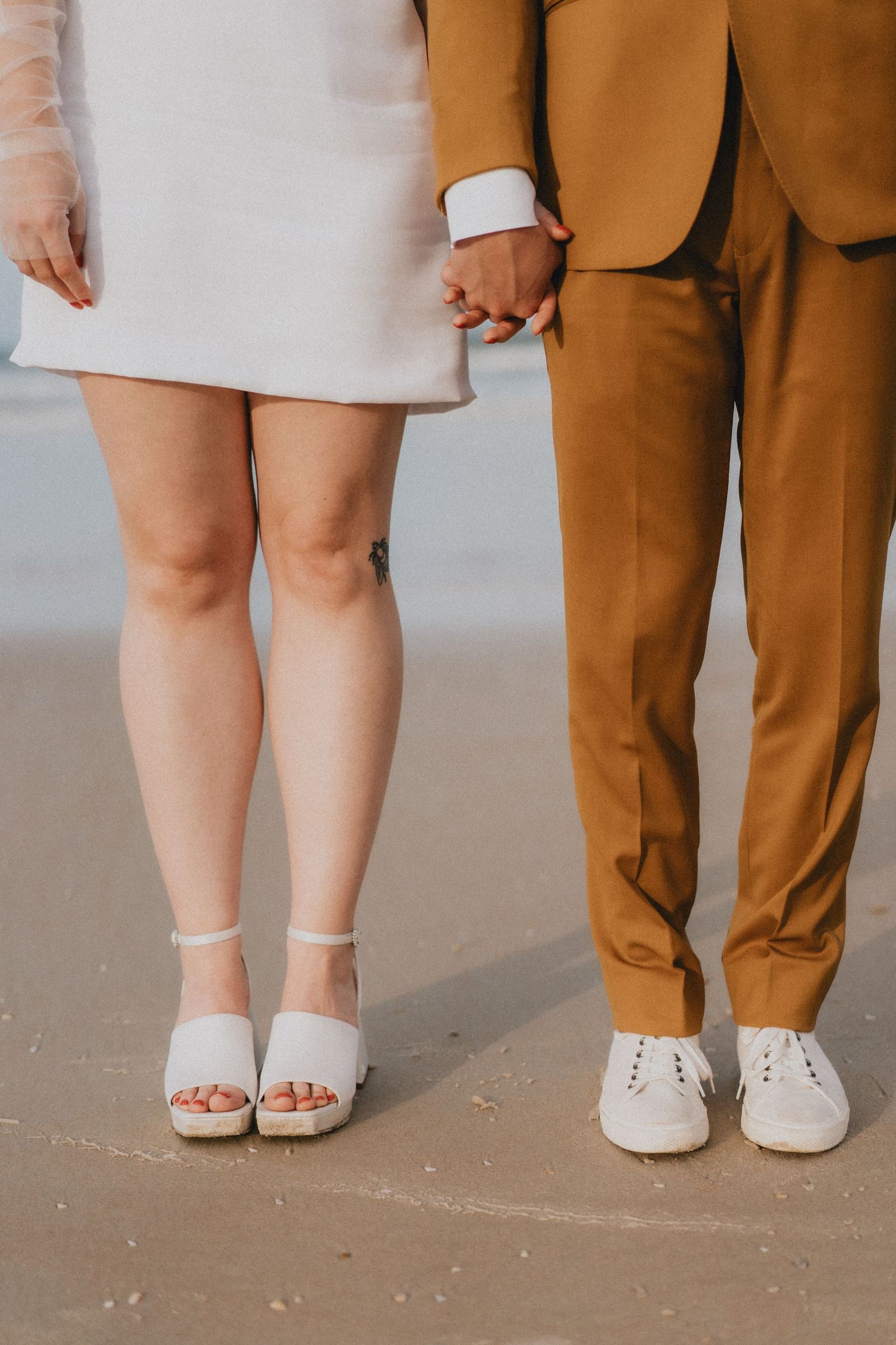 Close-up of bride and groom holding hands on beach, she in white heels, he in mustard suit and white shoes.