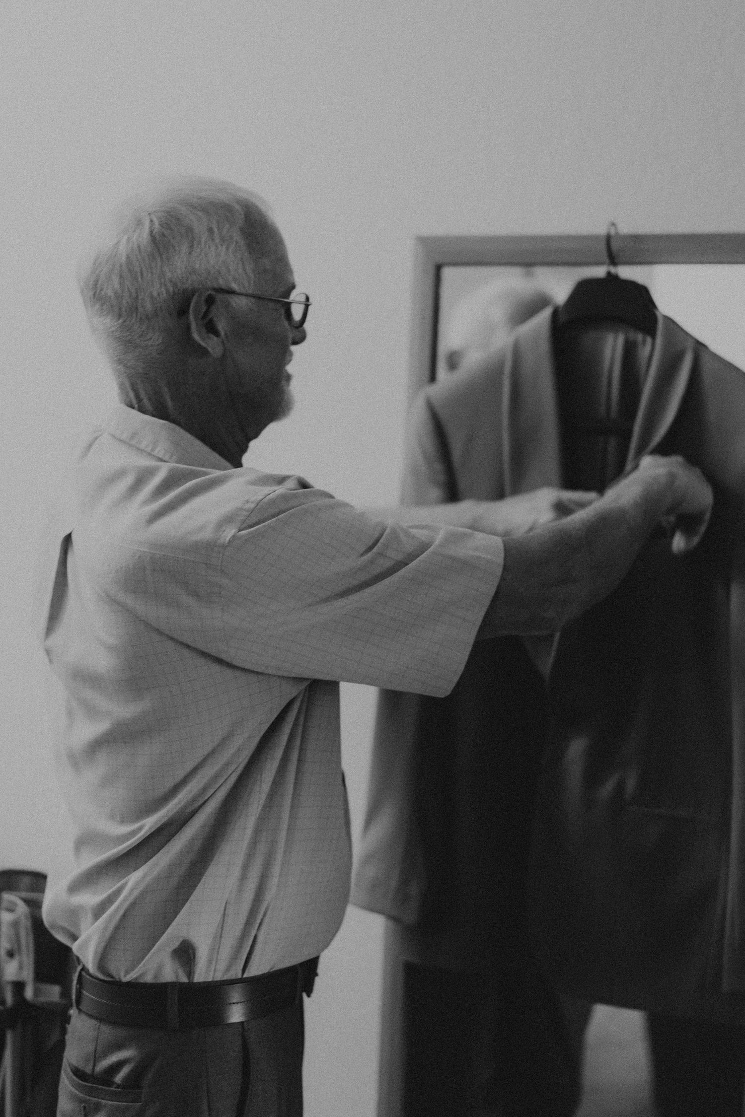 Elderly man in glasses selecting a suit jacket from a clothing rack in a black and white photo.