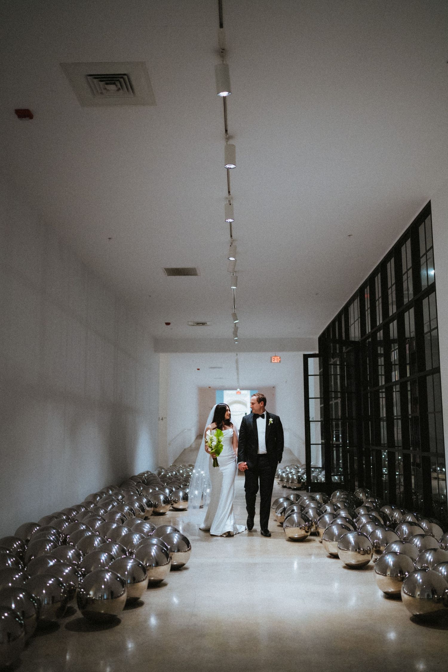 Bride and groom walk through a modern hallway lined with silver mirror balls and string lights at their elegant wedding venue.