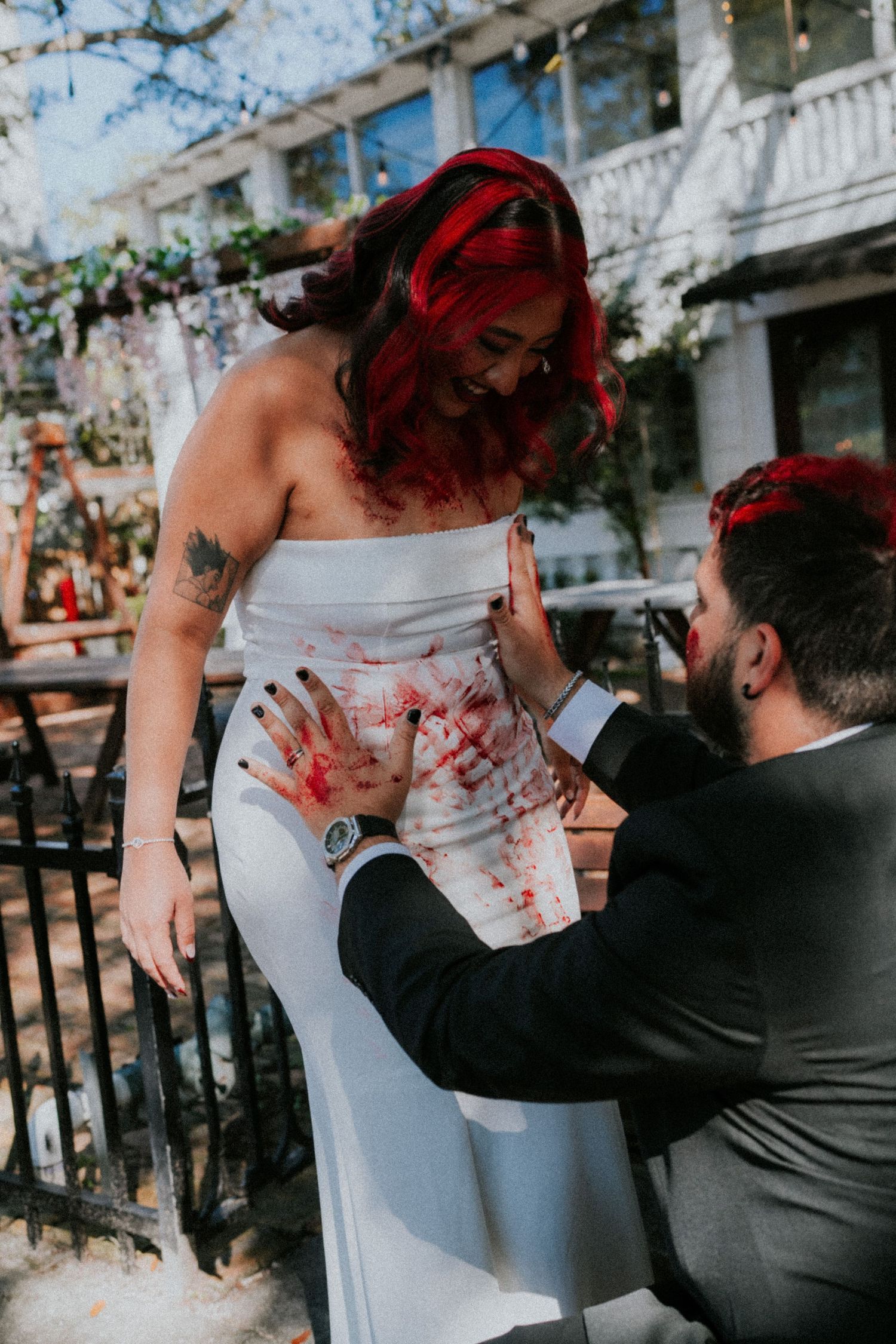 Bride with red hair laughs as groom kneels applying red paint to her white wedding dress outdoors.