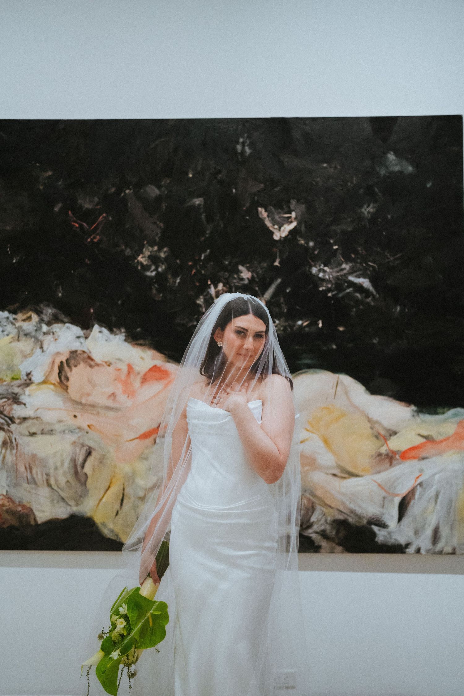Bride in white gown and veil holds greenery bouquet, posing before a dramatic dark abstract painting in a gallery.