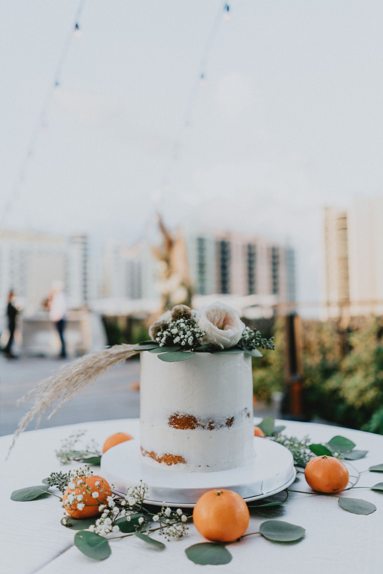 Elegant single-tier white semi-naked wedding cake topped with flowers, surrounded by oranges and eucalyptus on a white table.