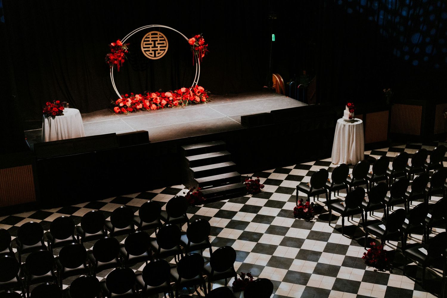 Elegant wedding ceremony setup with checkered floor, red floral arch, and rows of black chairs in a dark venue.
