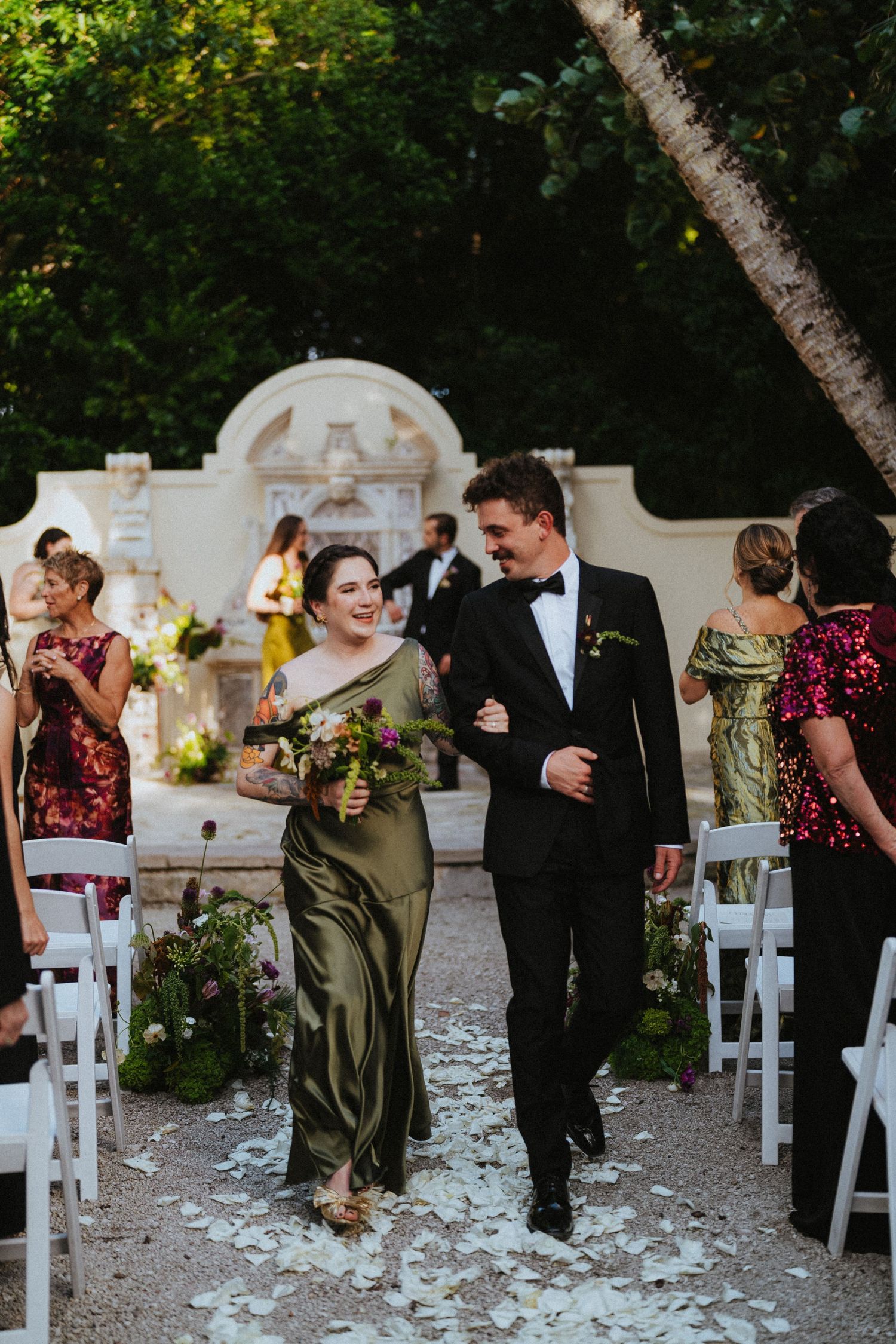 Smiling bridesmaid in olive green gown walks arm-in-arm with groomsman down petal-strewn outdoor wedding aisle.