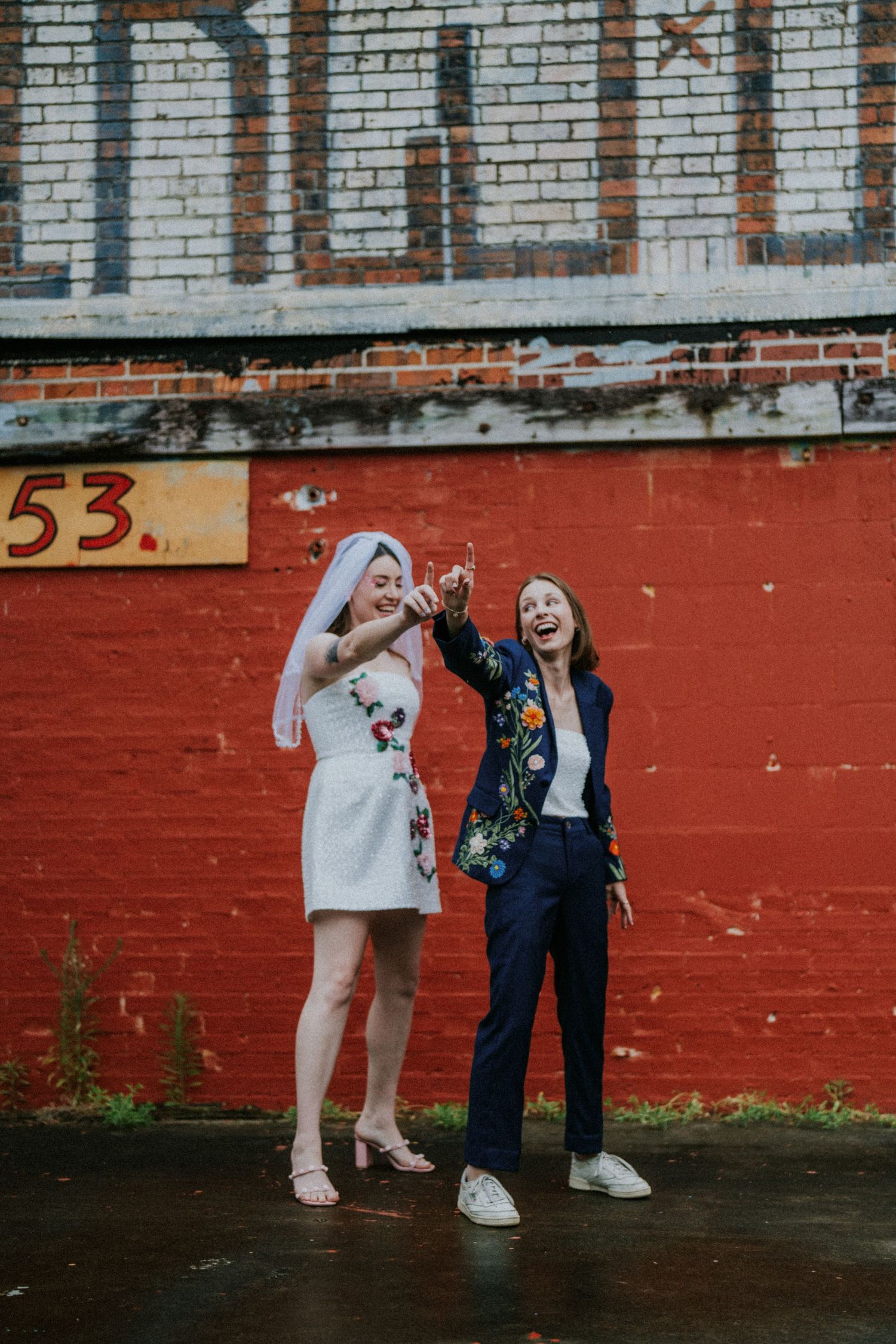 Two brides celebrate outdoors, one in floral mini dress with veil, other in navy floral suit, laughing by red brick wall.