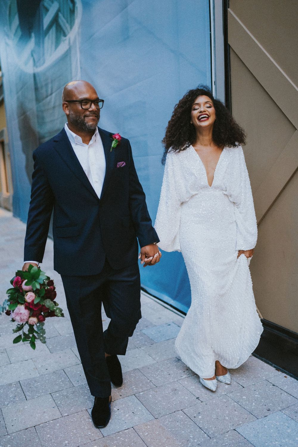 A couple walking together in formal attire, holding hands against a blue wall background.