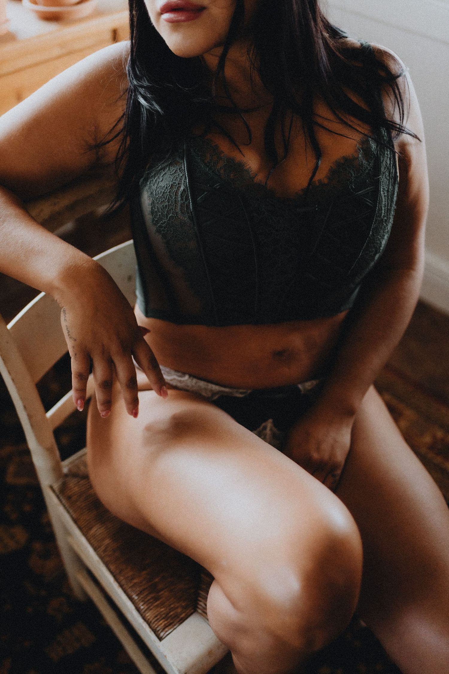 Person wearing black lingerie sits on wooden chair in intimate boudoir setting.