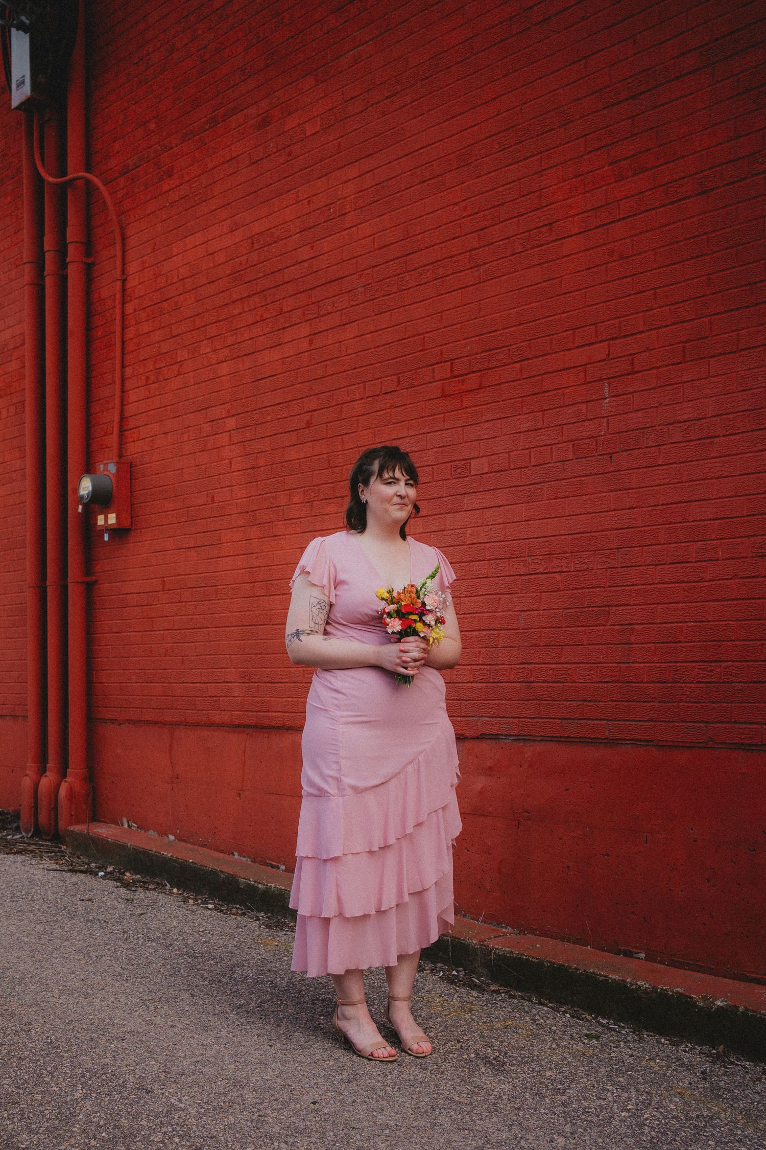 Woman in pink tiered dress holding colorful bouquet, smiling against red brick wall backdrop.