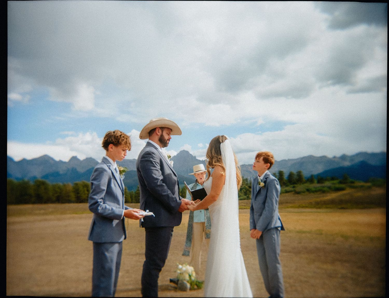 Outdoor mountain wedding ceremony with bride, groom in cowboy hat, and two ring bearers under cloudy skies.