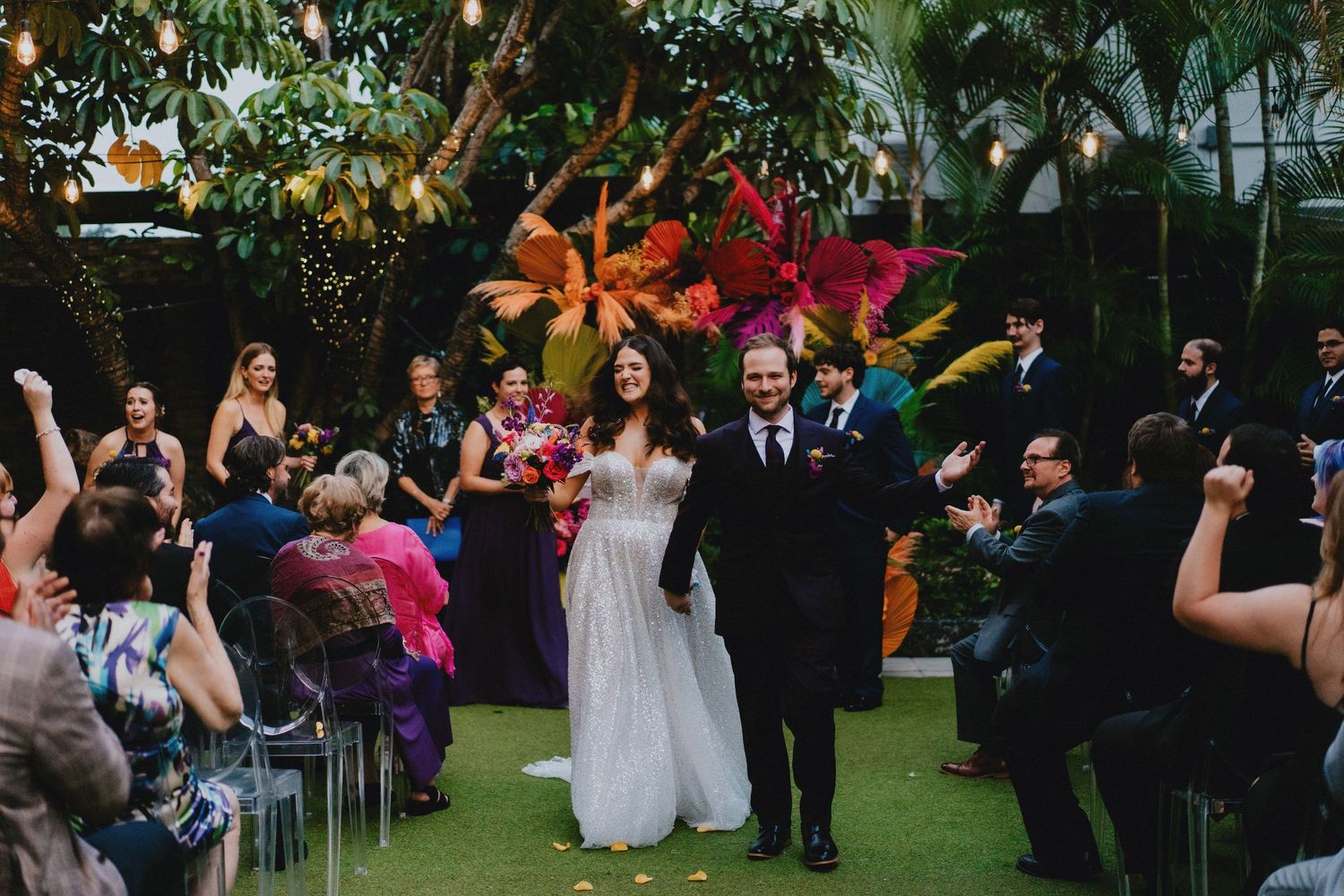 Newlywed couple walks down the aisle at a vibrant outdoor wedding ceremony with colorful floral backdrop.