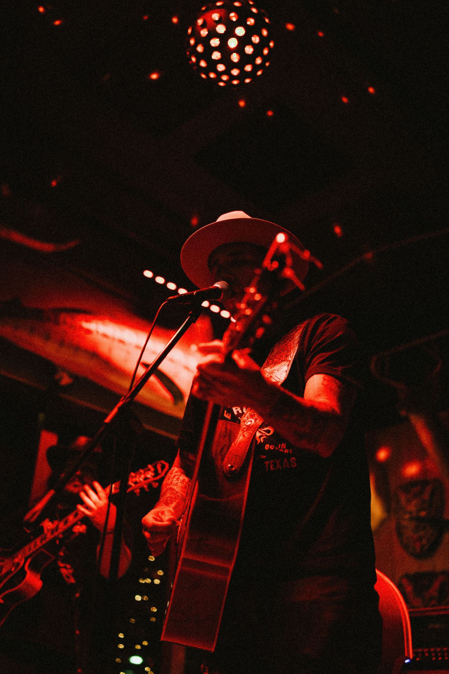 Musician in white hat plays acoustic guitar on dimly lit red-toned stage with disco ball overhead.