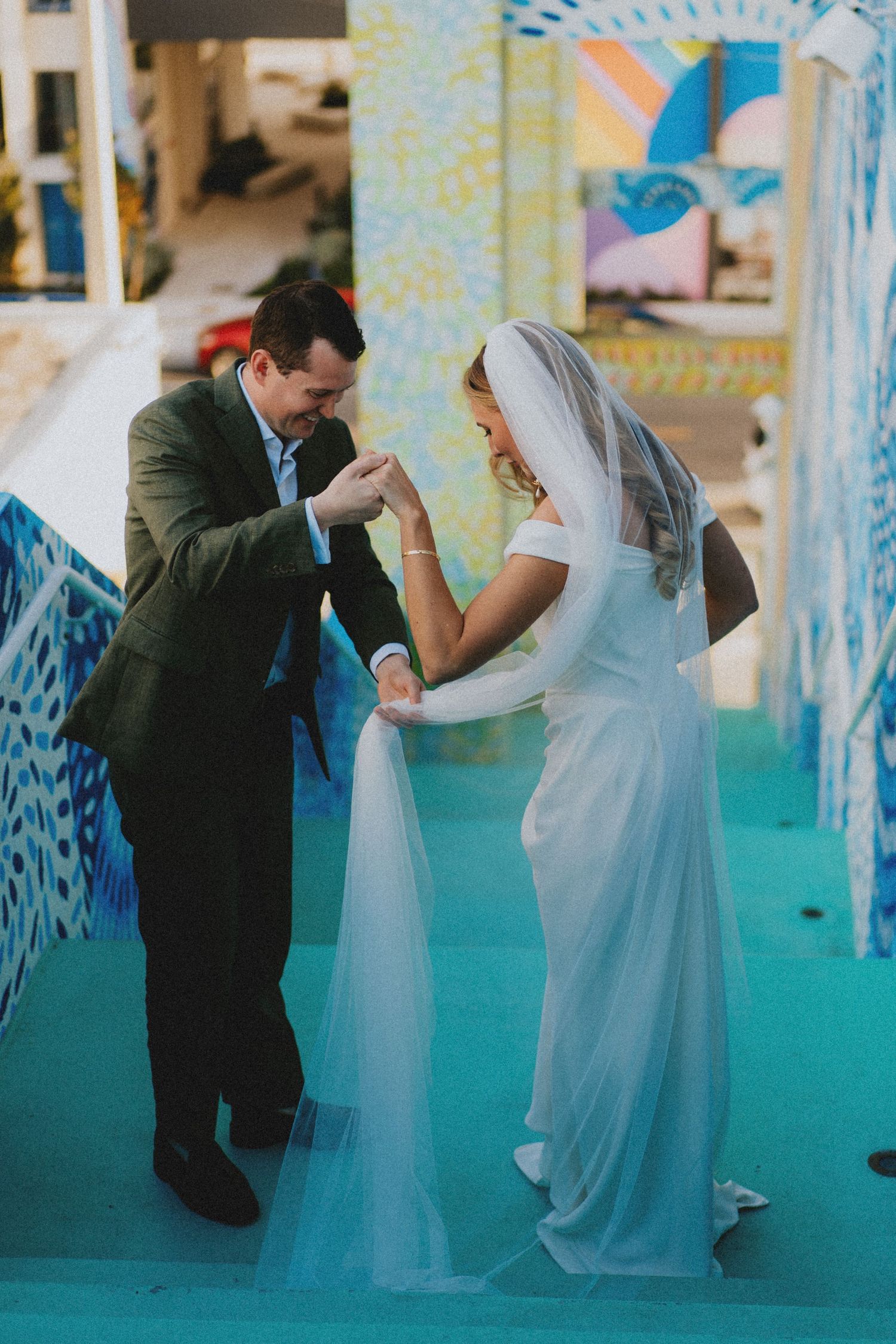 Groom helps bride with her veil on colorful blue steps during wedding ceremony.