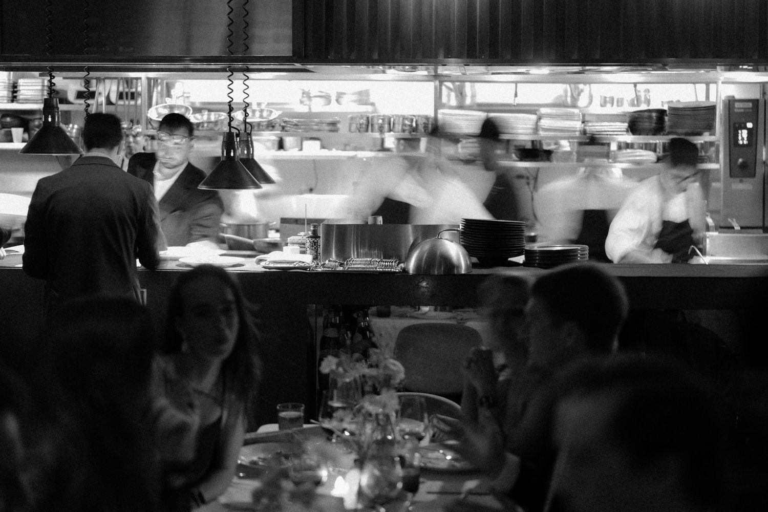 Black and white photo of a busy restaurant kitchen with blurred chefs cooking while diners sit at candlelit tables in foreground.