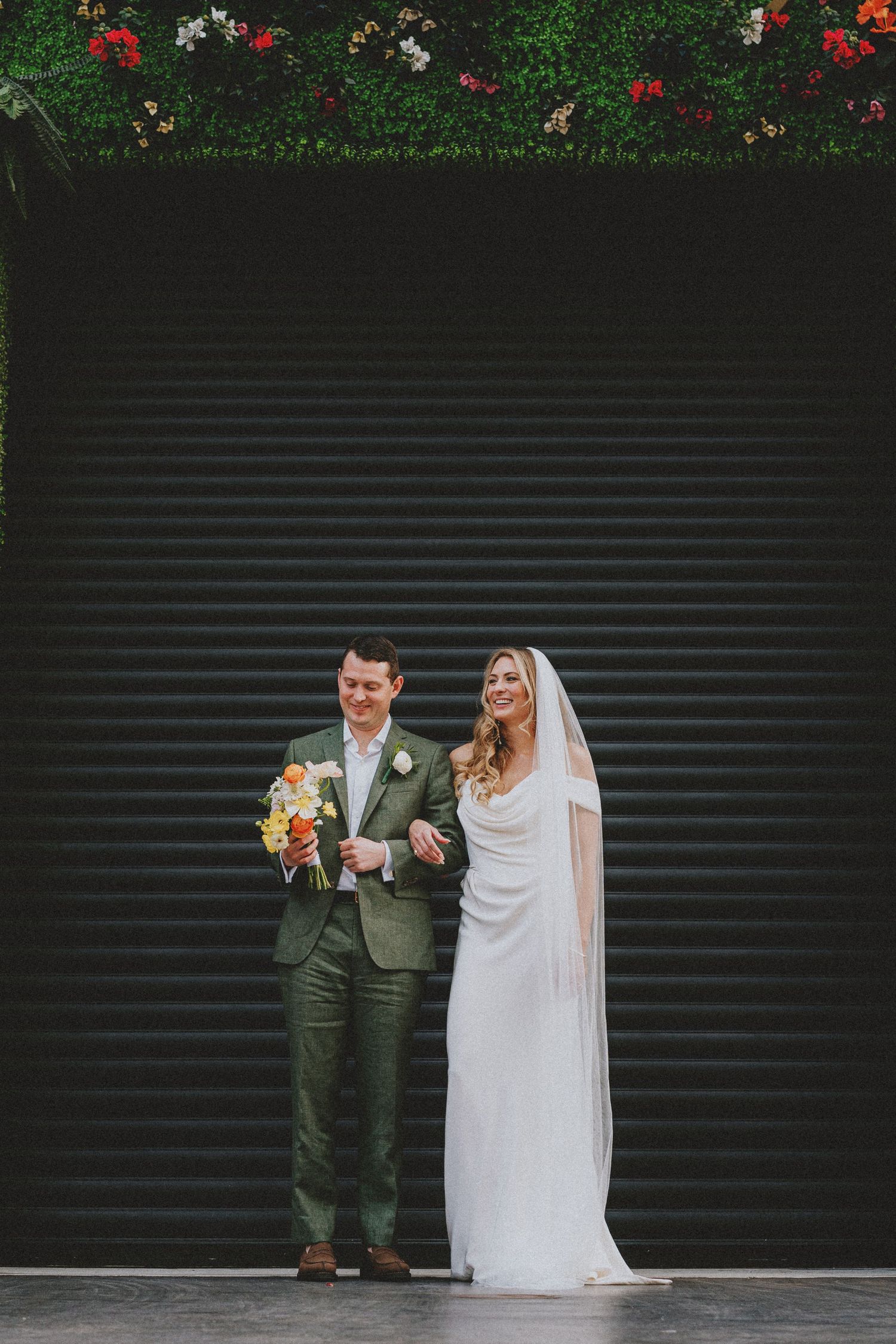 Bride in white gown and veil stands with groom in green suit holding colorful bouquet against dark corrugated wall.