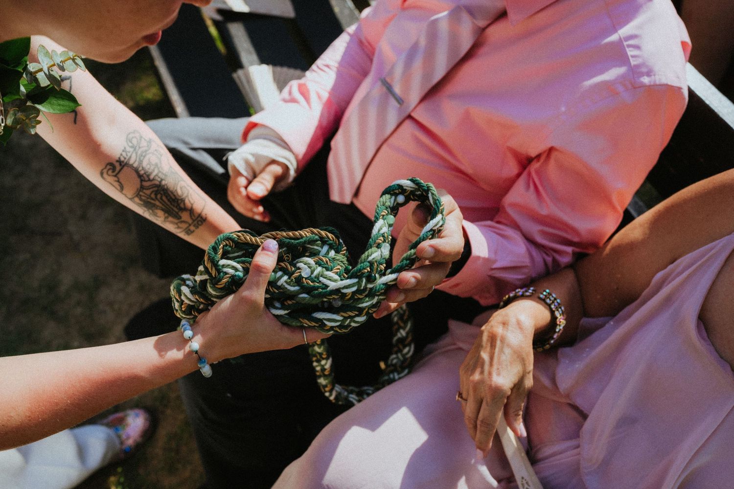 Close-up of hands holding a green floral fabric accessory, with tattoos and bracelets visible, outdoors in sunlight.
