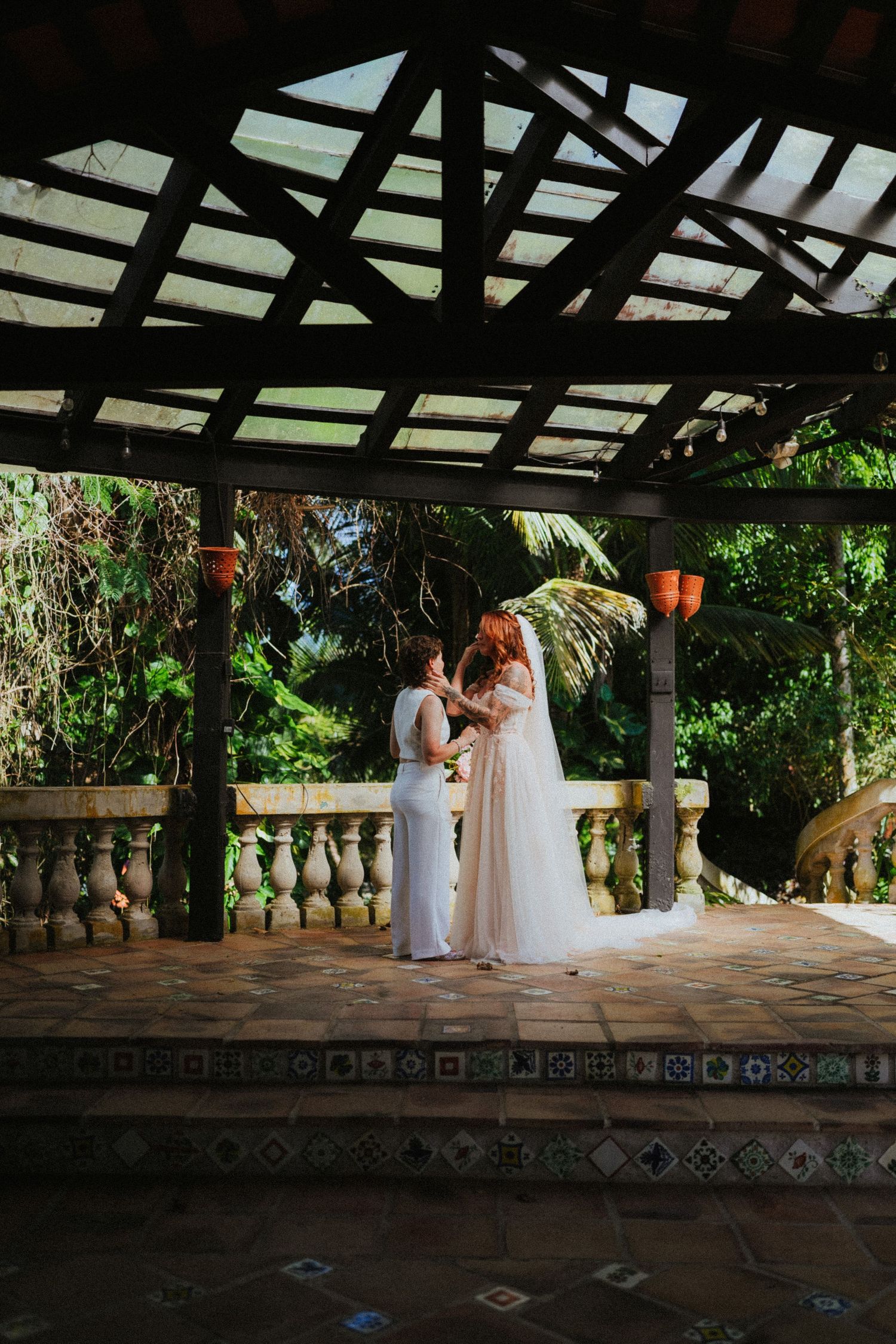 Two brides share a tender moment under a rustic pergola surrounded by lush tropical greenery at an outdoor wedding.