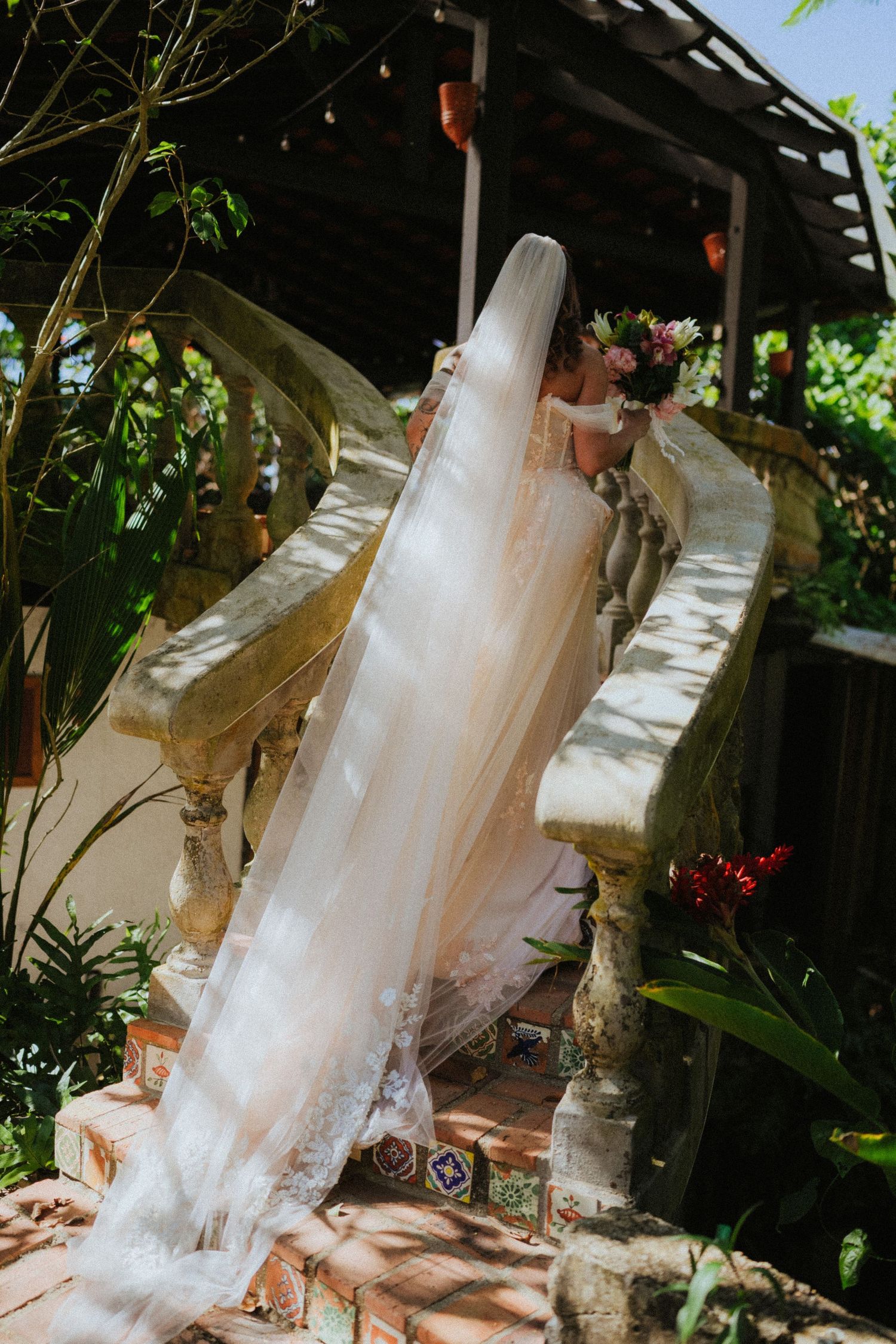 Bride ascending stone stairs in a flowing white gown with long lace veil, holding a colorful bouquet in a lush garden.