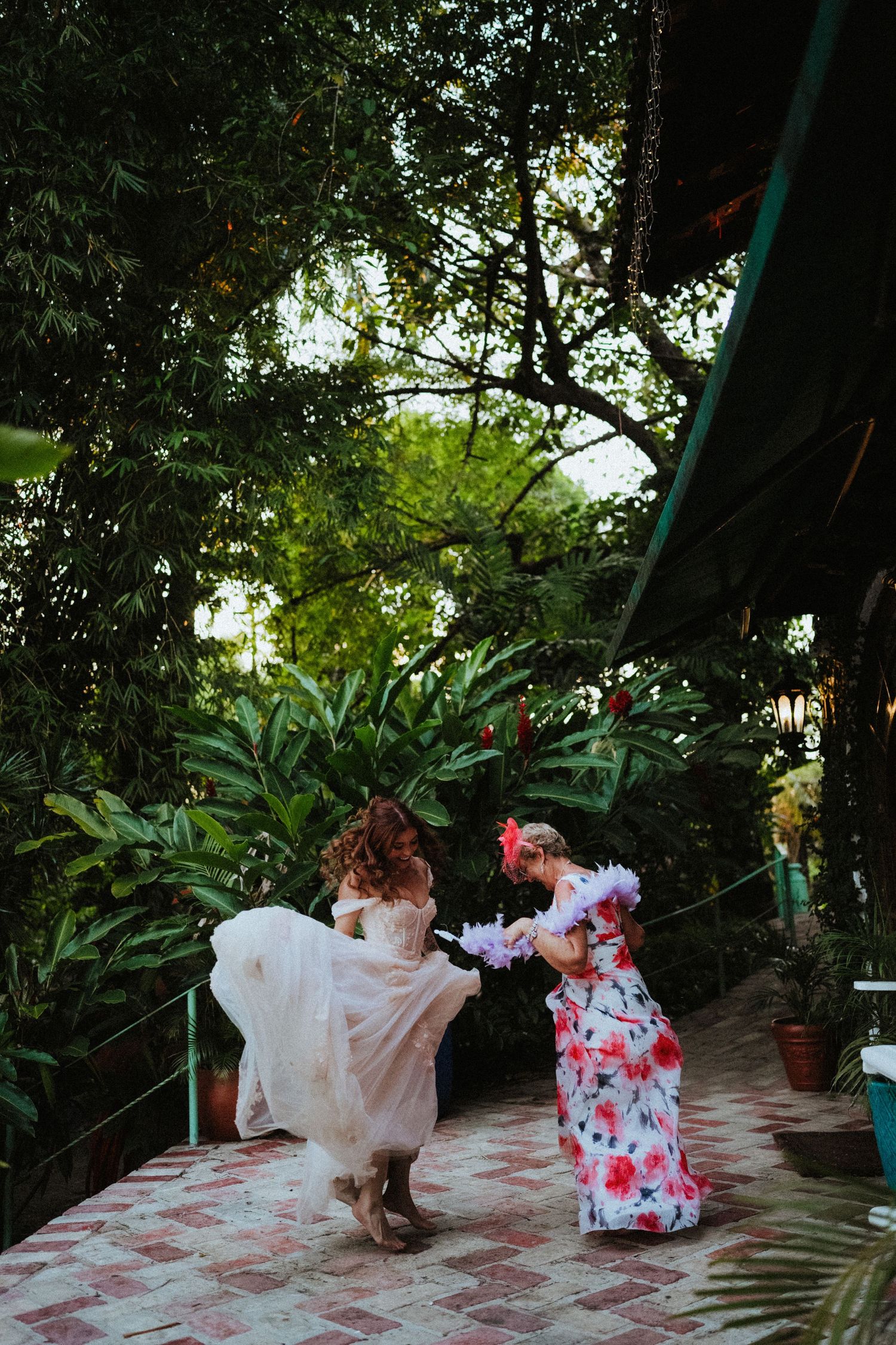 Two women in elegant dresses dance joyfully on a brick patio surrounded by lush tropical greenery.