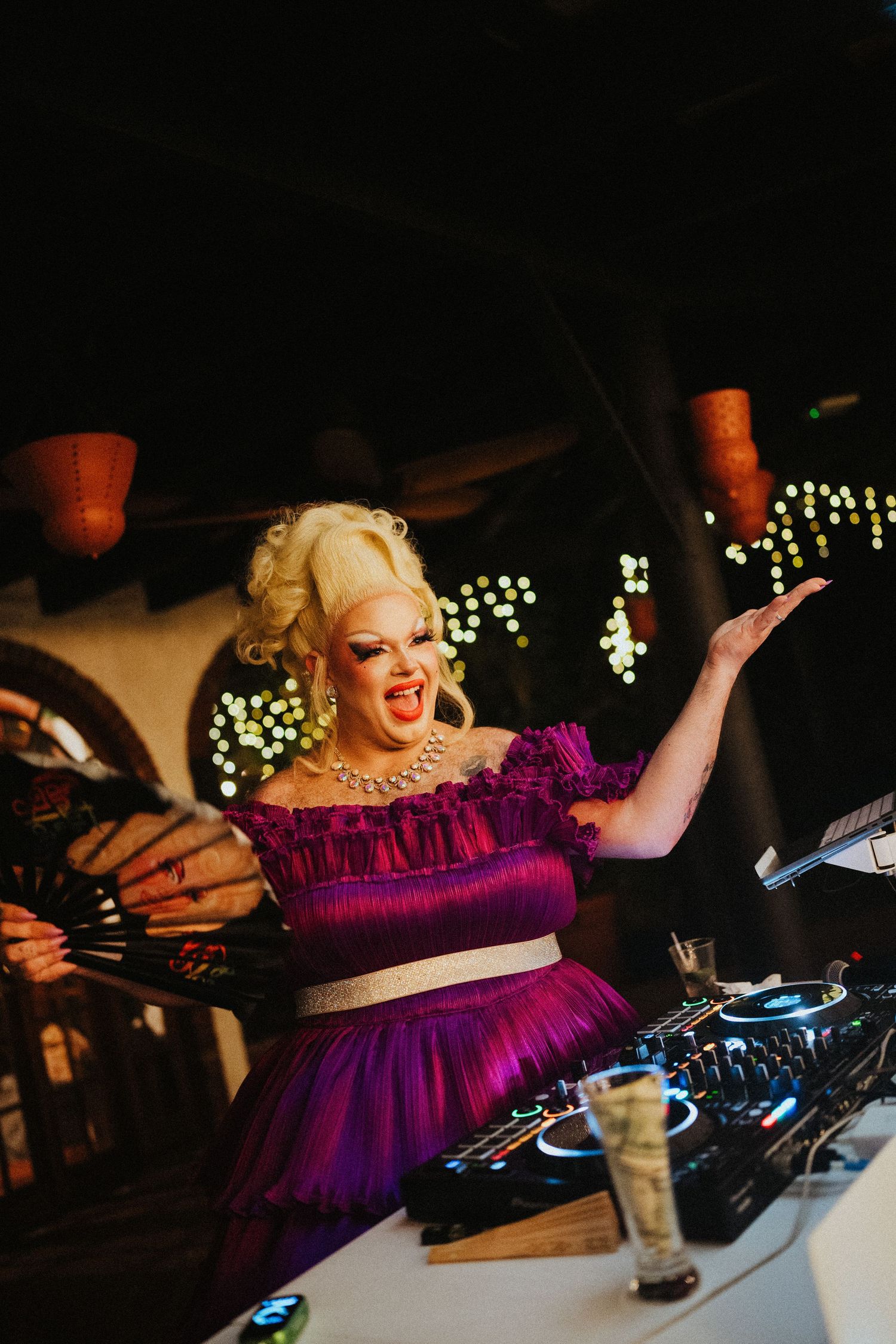 Drag queen in purple ruffled dress and blonde wig performing at DJ booth with hands raised at vibrant nightclub event.