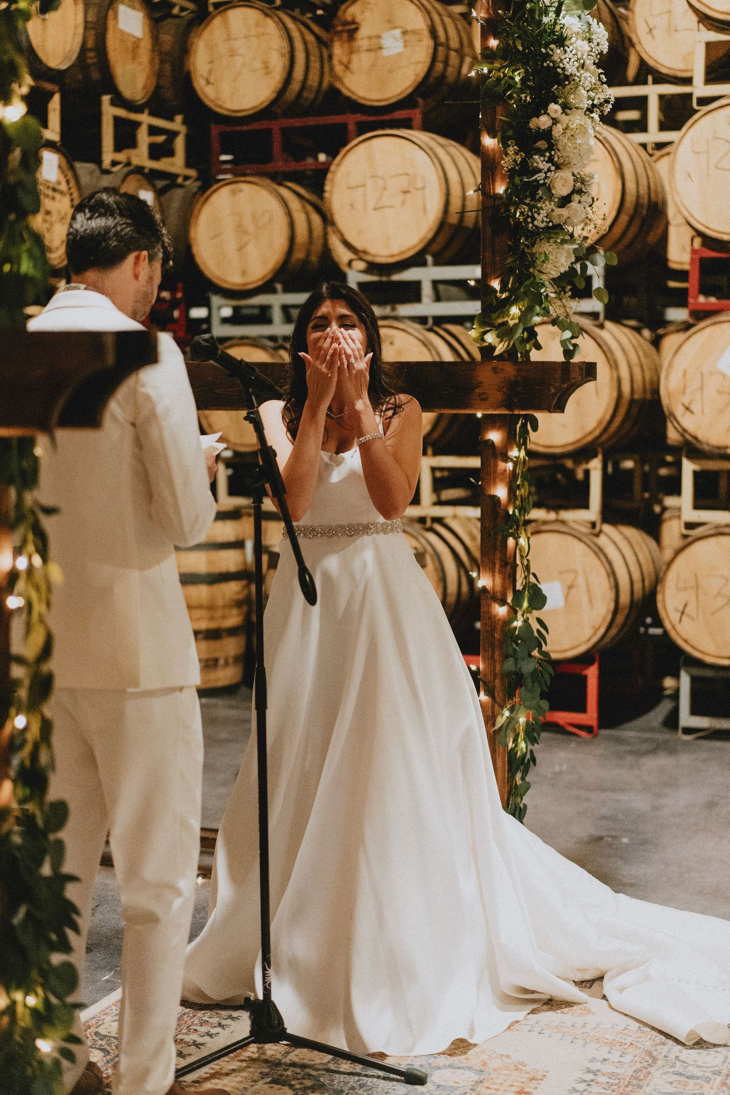 Bride covers her mouth in tears during emotional winery wedding ceremony surrounded by oak barrels and floral arch.