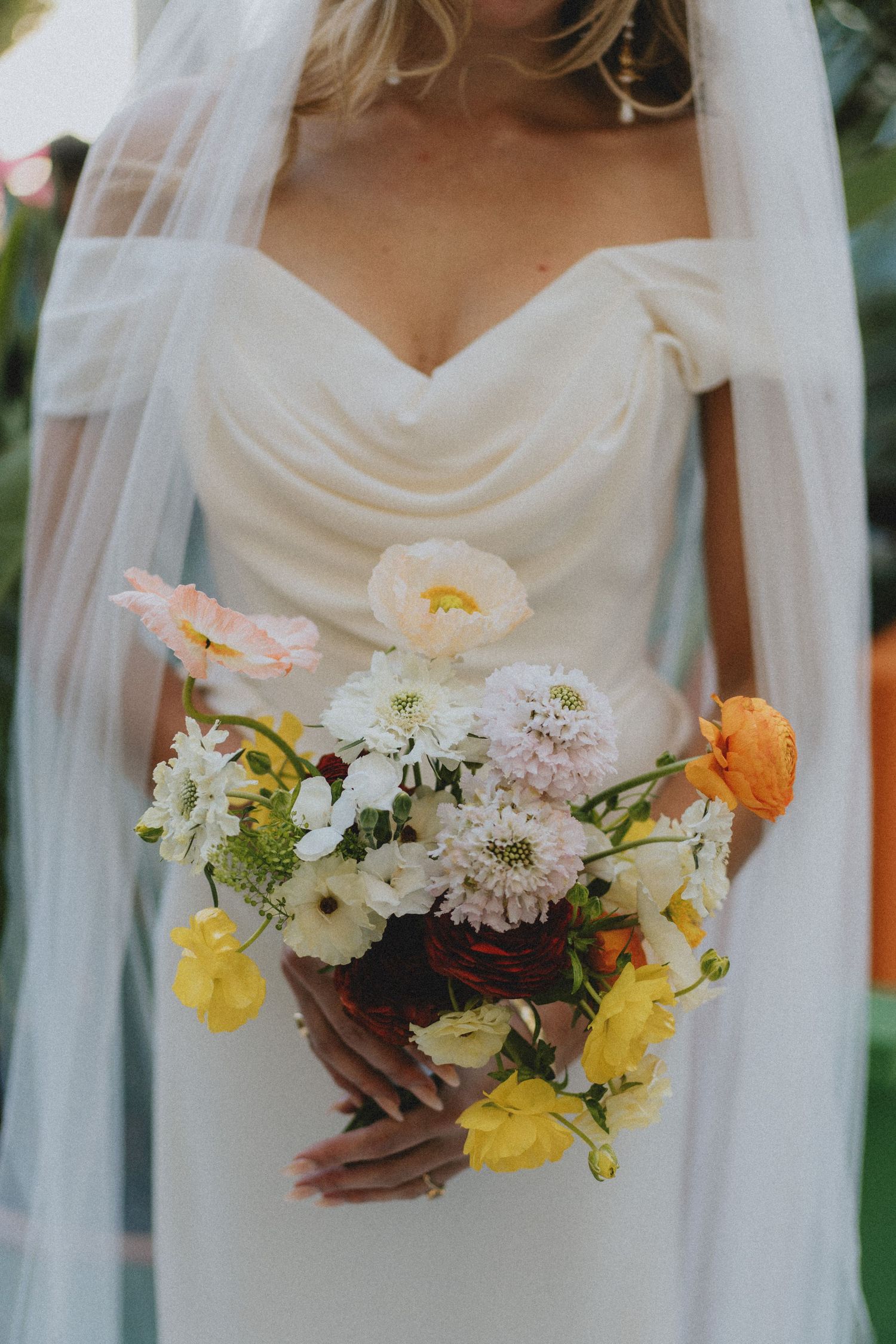 Bride holding a colorful wedding bouquet with pink, orange, and yellow flowers in a white off-shoulder gown.