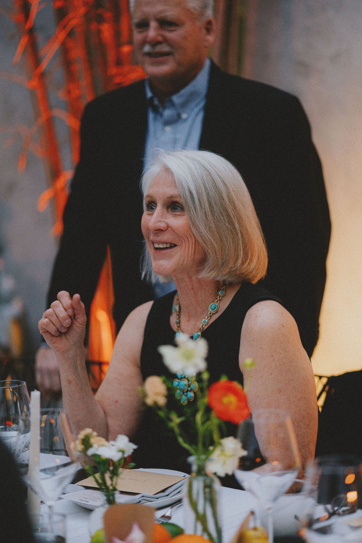 Elegant older woman with gray bob smiling at dinner party wearing turquoise necklace and black dress.
