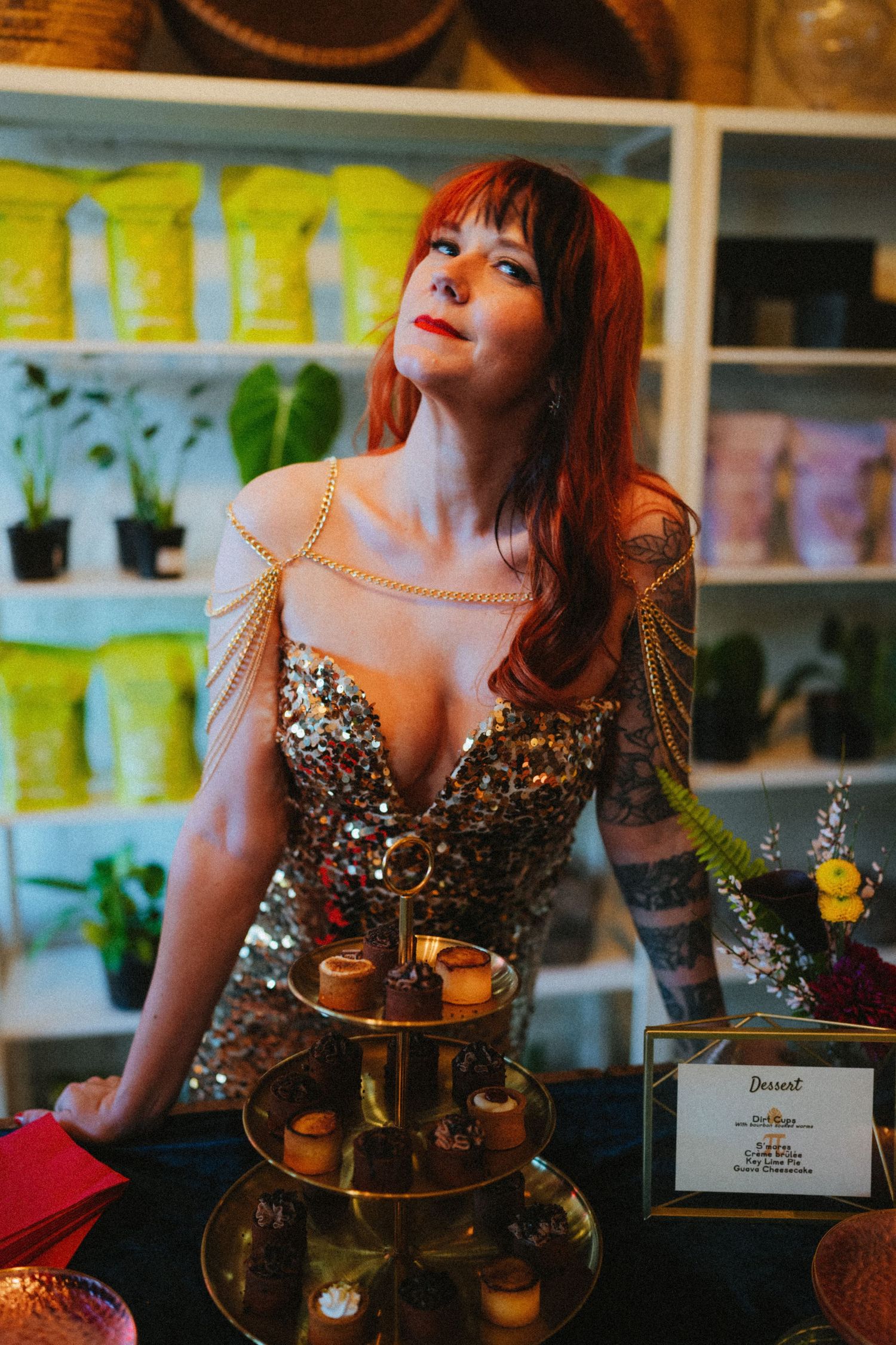 Red-haired woman in sequined gown with gold chain jewelry stands beside tiered dessert display in boutique shop.