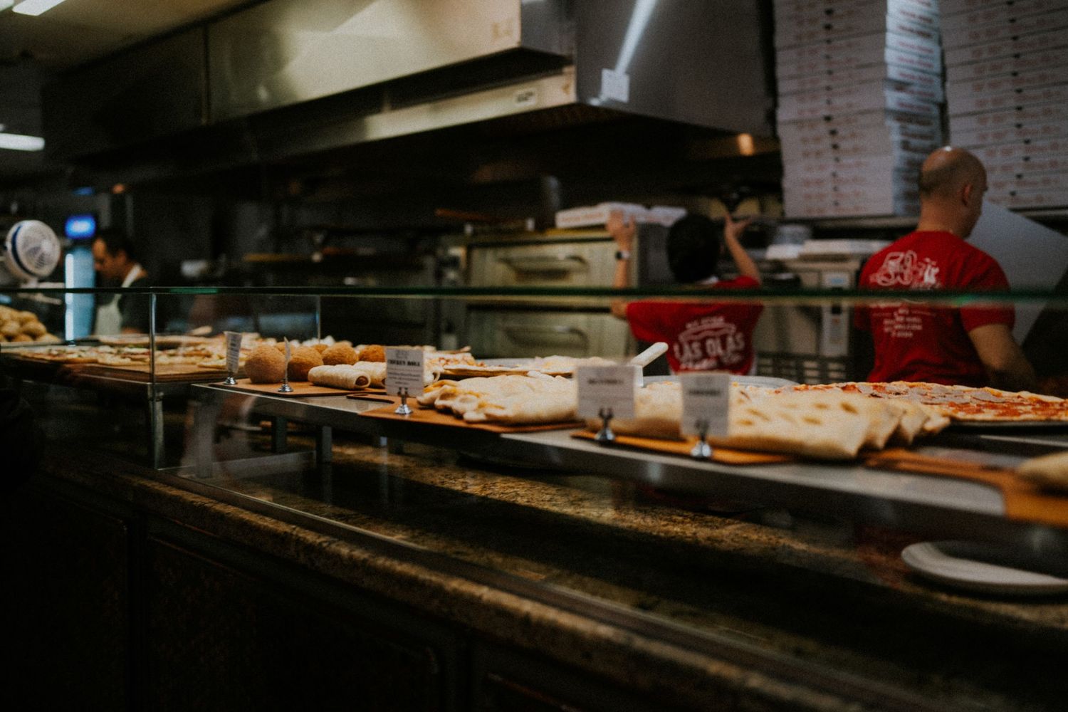 Pizza restaurant kitchen with staff in red shirts preparing food on a stainless steel counter with ovens in background.