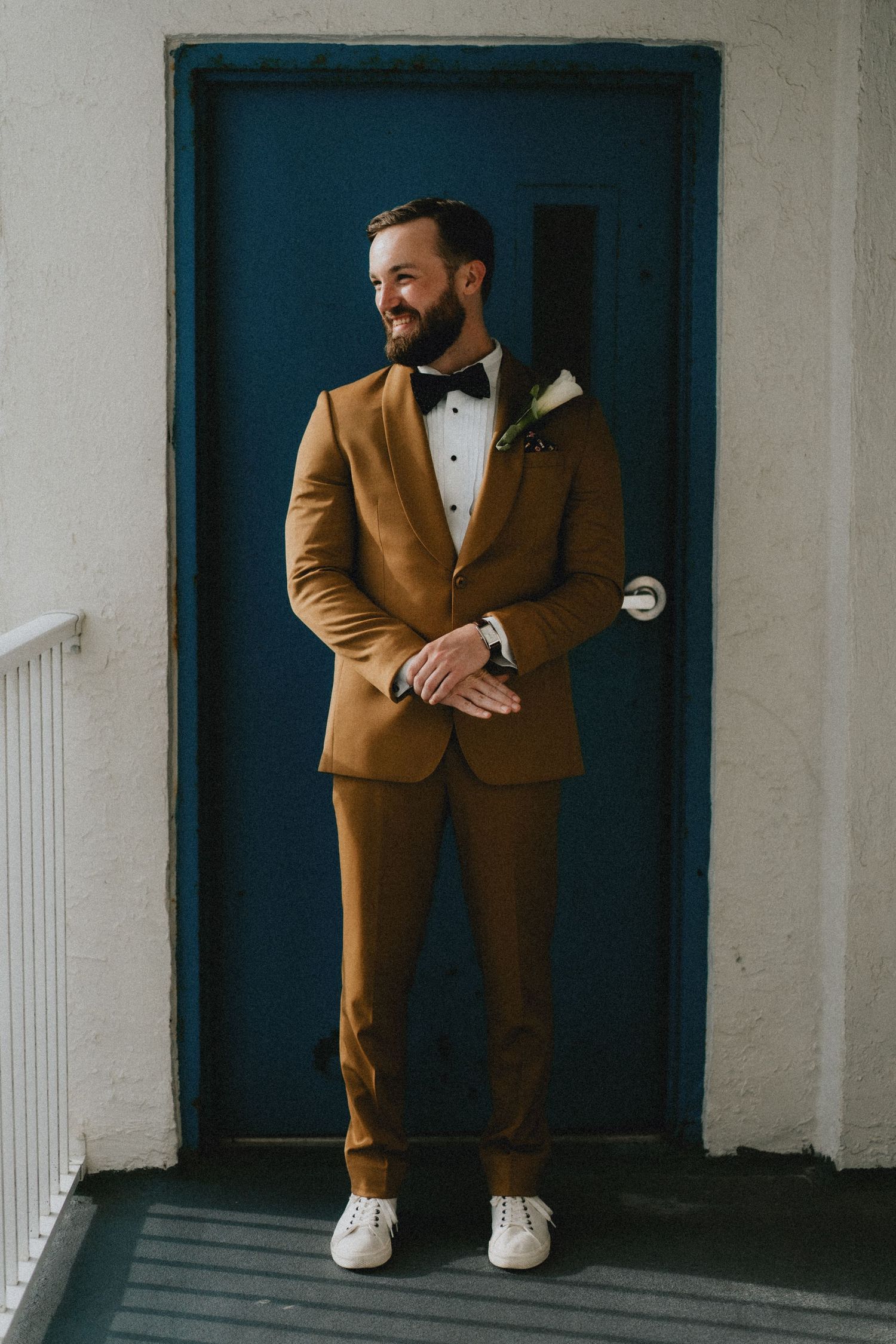 Groom in mustard suit with black bow tie and boutonniere stands smiling before a teal door.