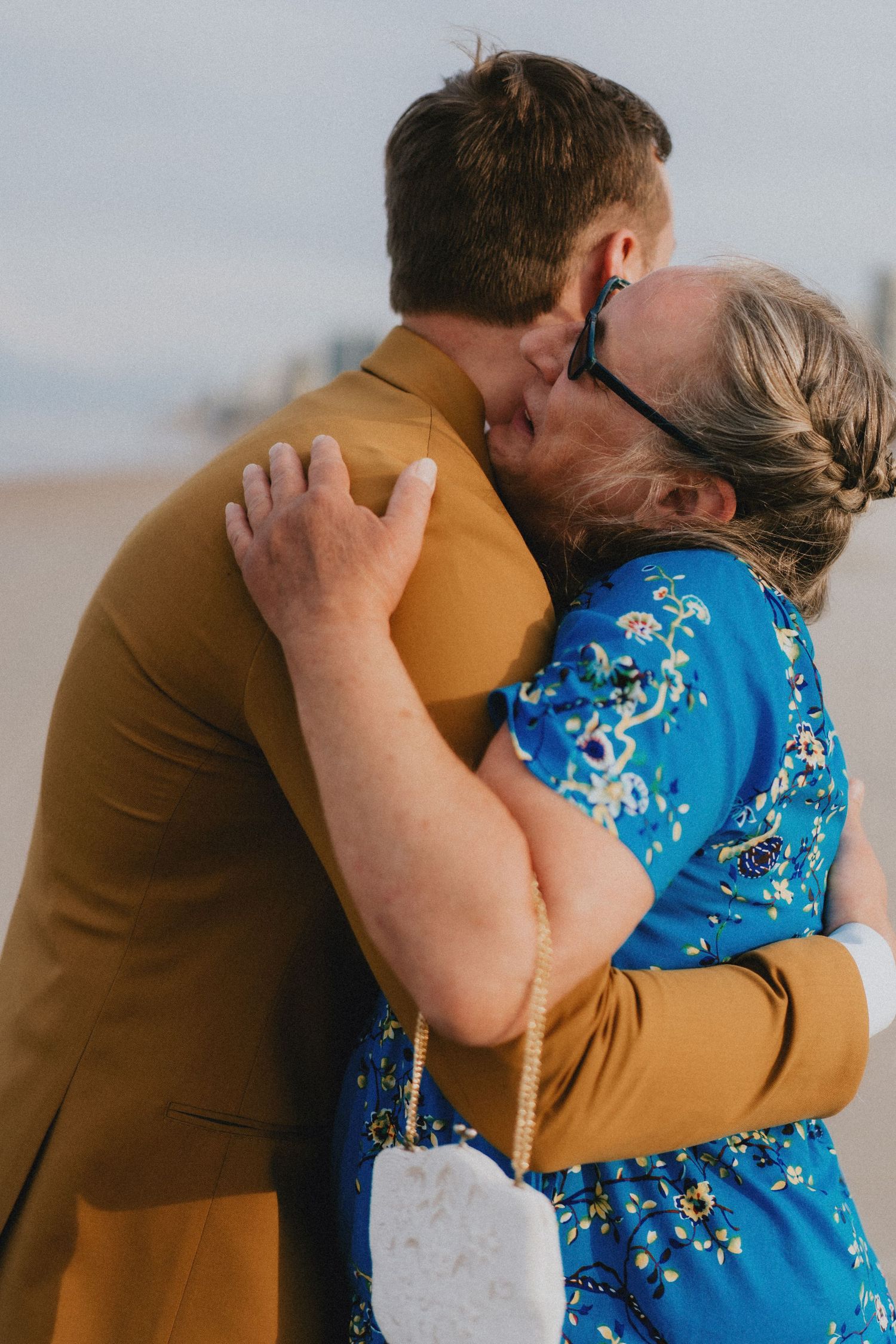 A man in a tan suit shares a tender kiss with an older woman in a blue floral dress on a beach.