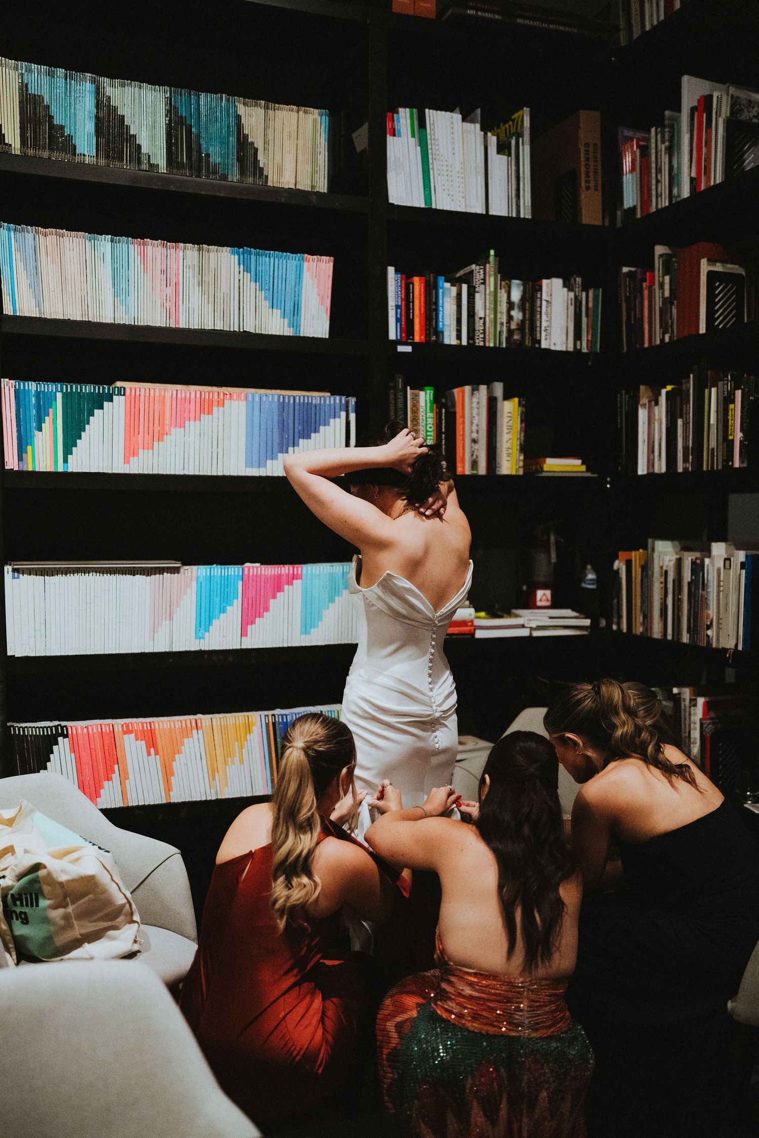 Bride in white dress stands before colorful bookshelves as bridesmaids in formal gowns help with her wedding dress.