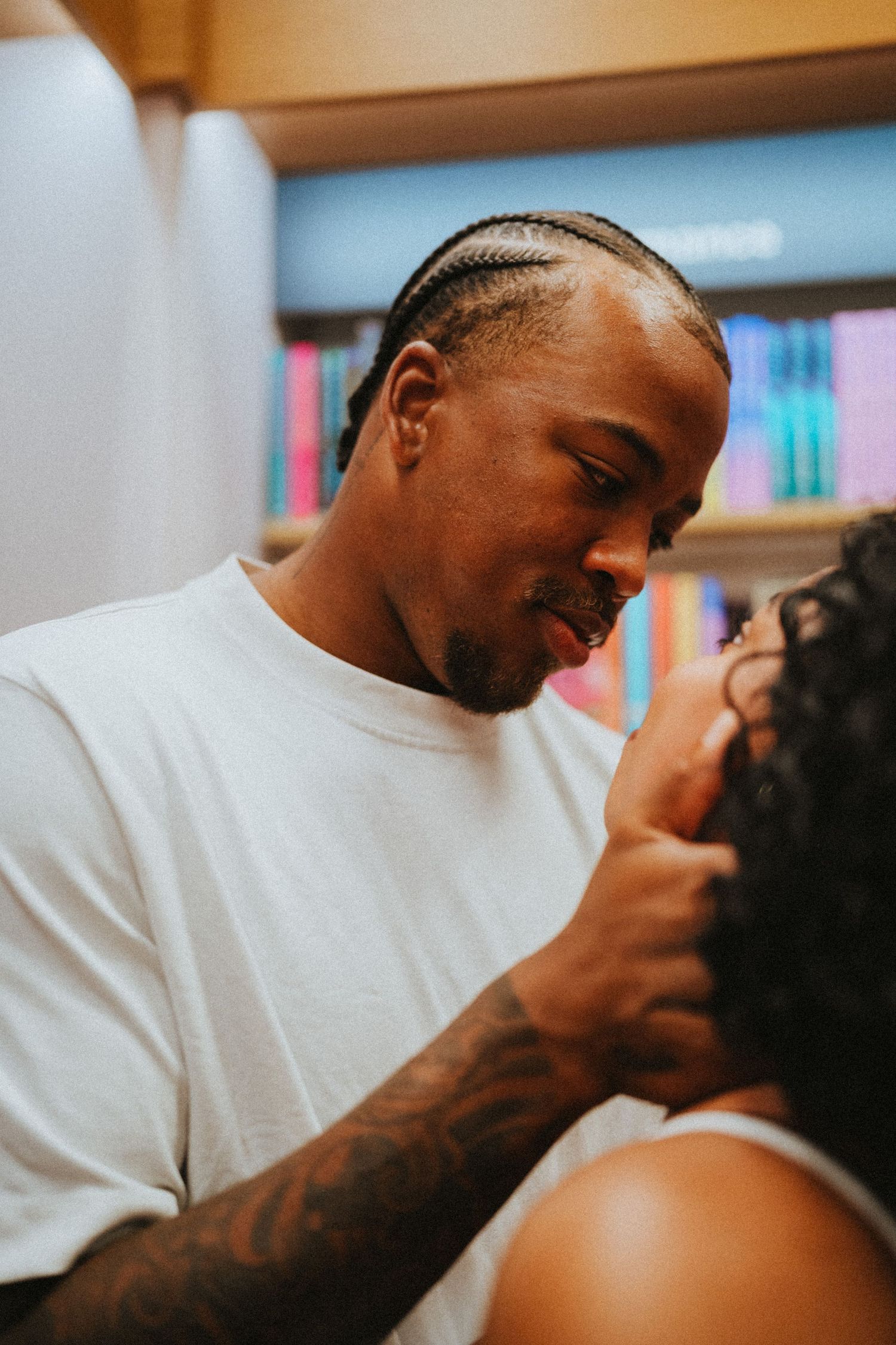 Tattooed man gazing lovingly at woman in front of colorful bookshelves.