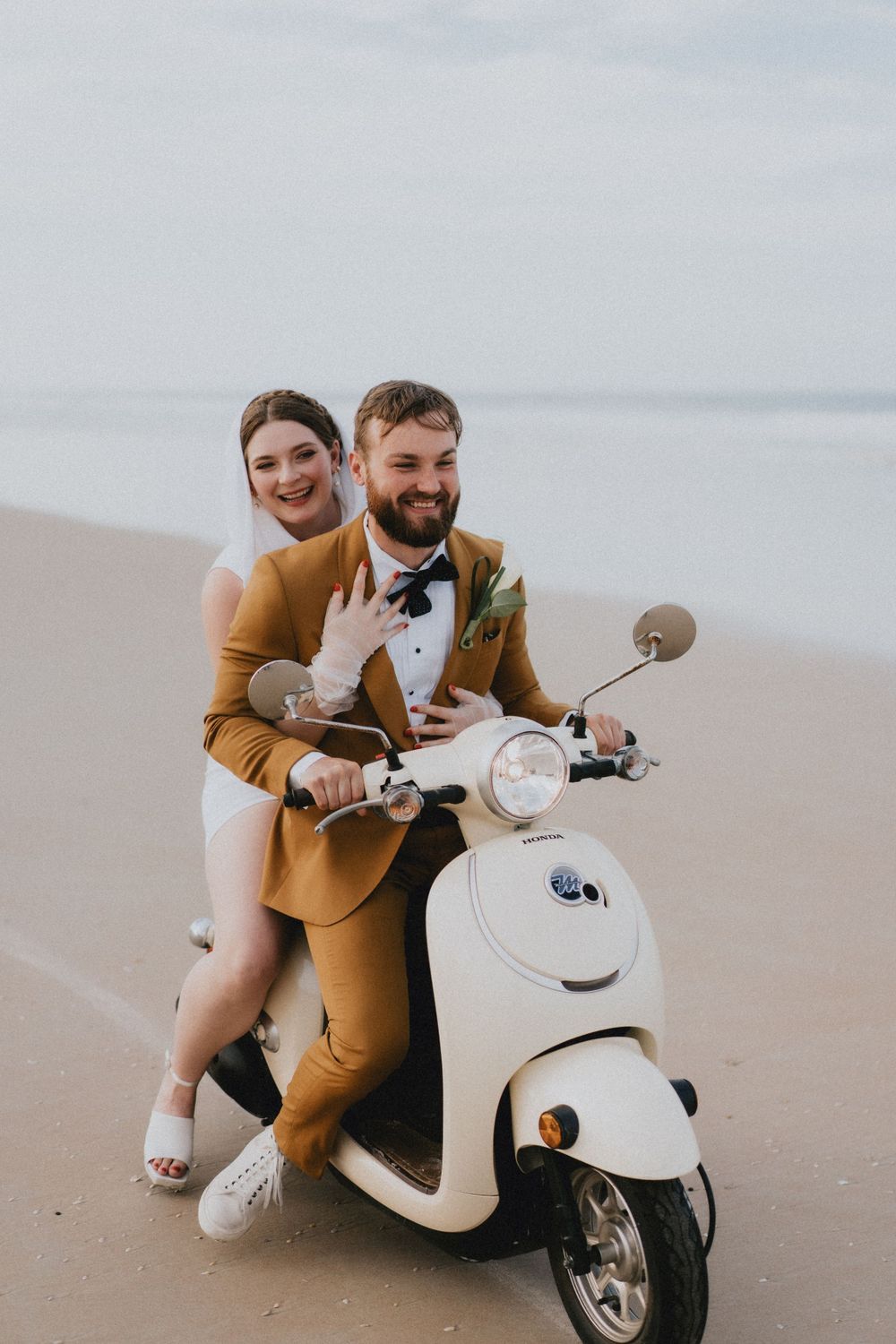 Playful couple poses with a white Vespa scooter on a sandy beach.