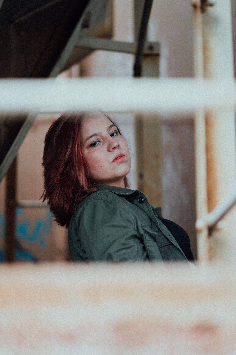 Moody portrait through window blinds showing someone in a green jacket looking upward.