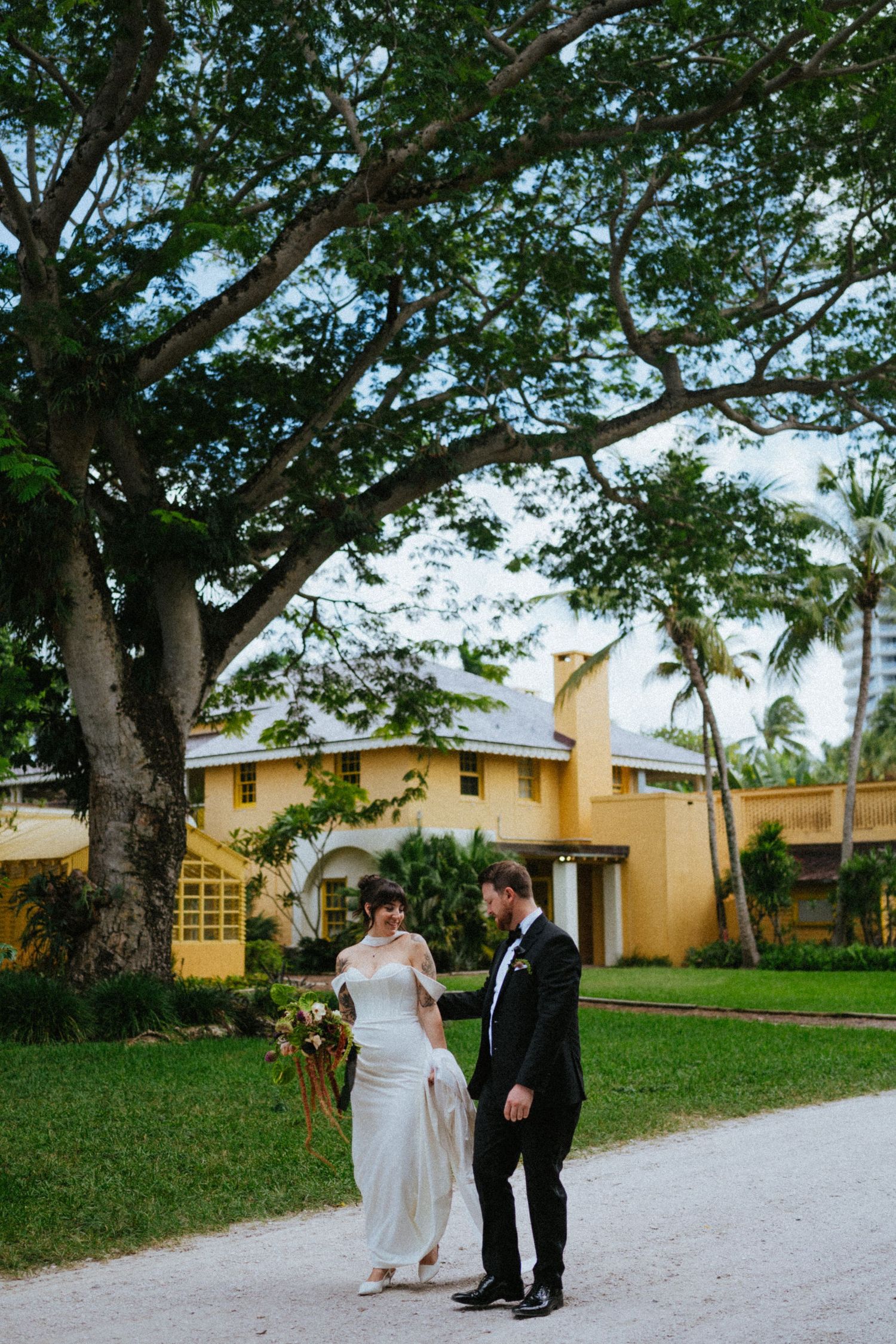 Bride and groom walk hand in hand on a garden path beneath a large tree at a yellow estate venue.