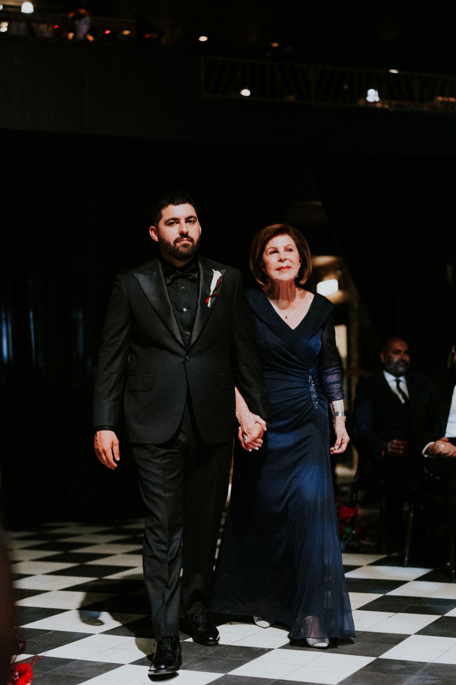 Groom in black tuxedo escorts elegant woman in navy gown across checkered dance floor at formal wedding reception.