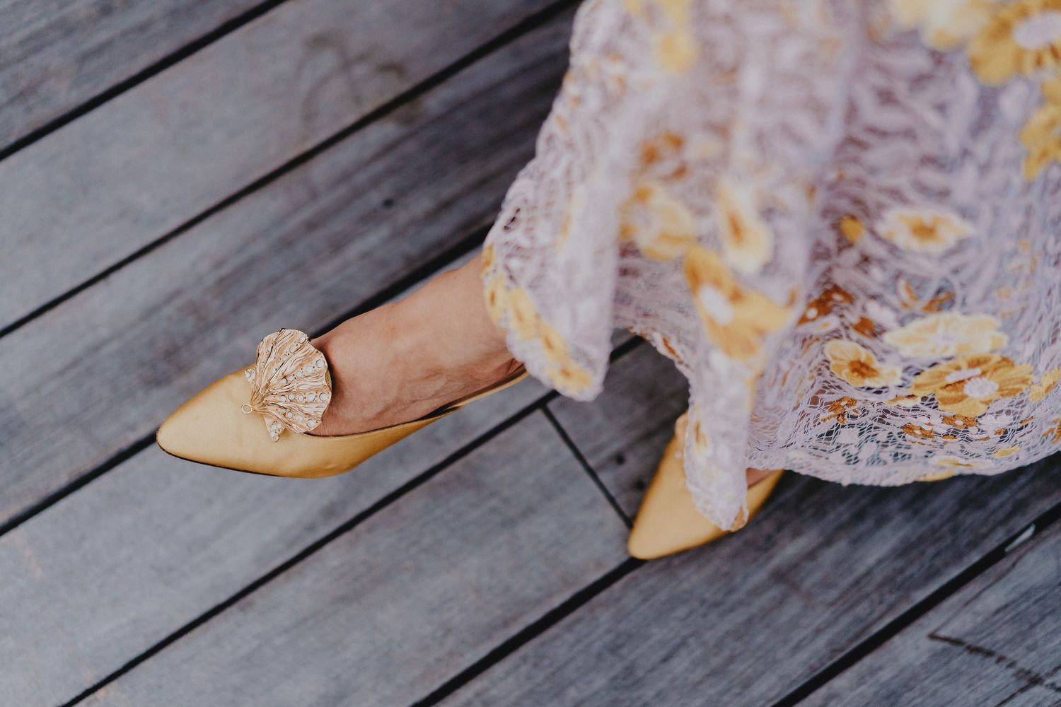 Yellow flat shoes with floral embellishment worn with a lace floral dress on a wooden deck.