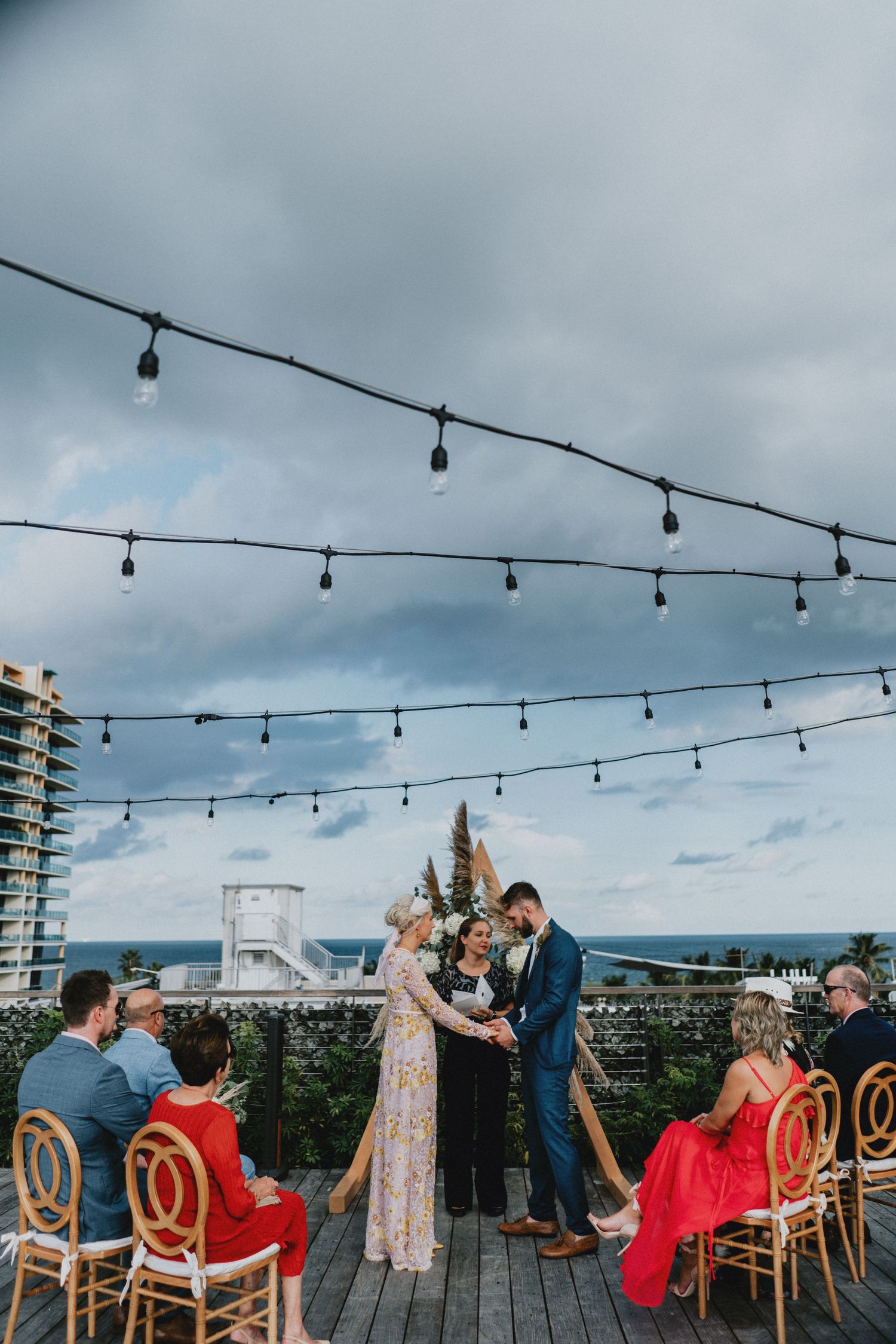 Couple exchanges vows on a rooftop terrace with string lights, ocean views, and guests seated in wooden chairs under cloudy skies.