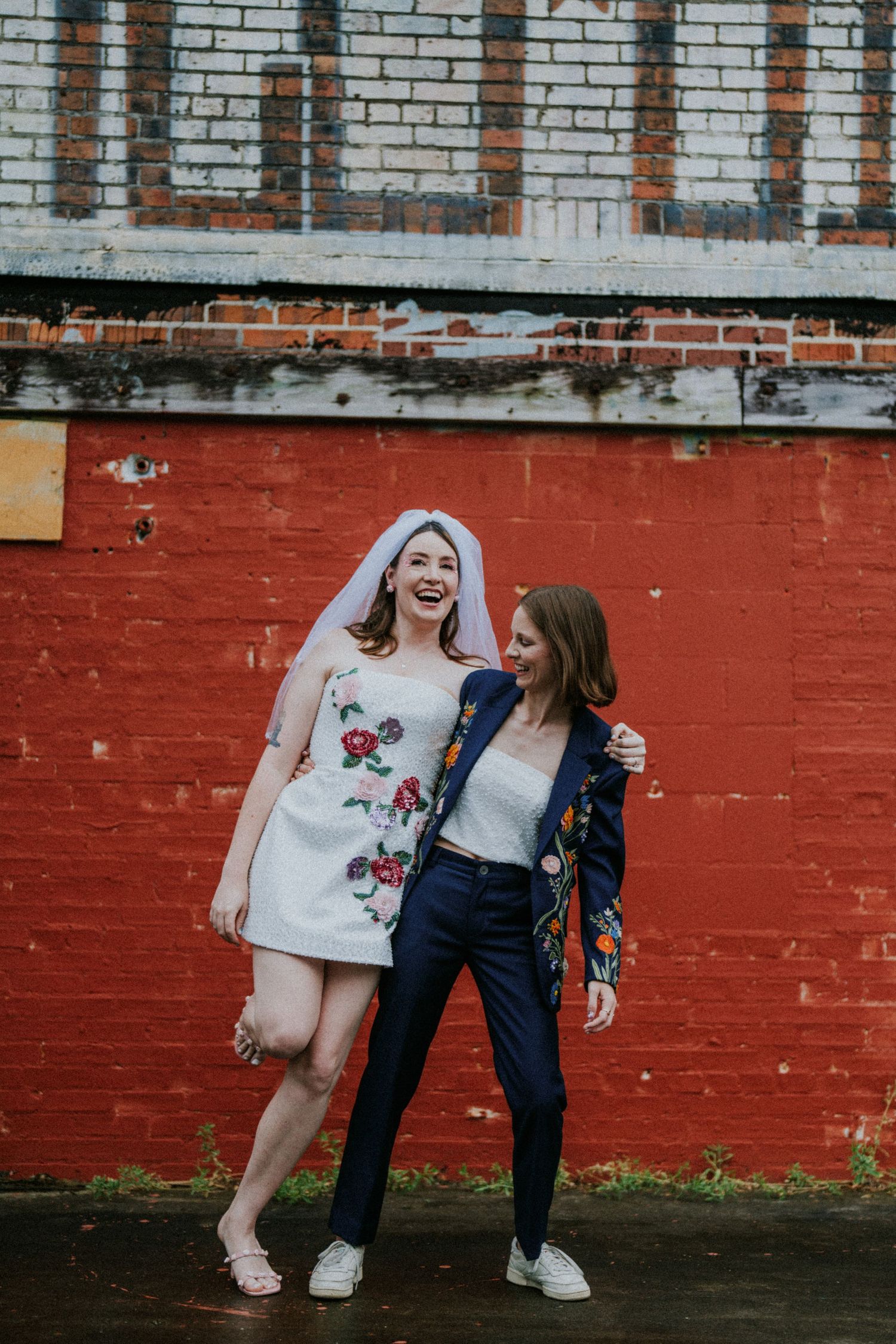 Two brides pose happily by red brick wall, one in floral mini dress with veil, other in navy embroidered blazer.