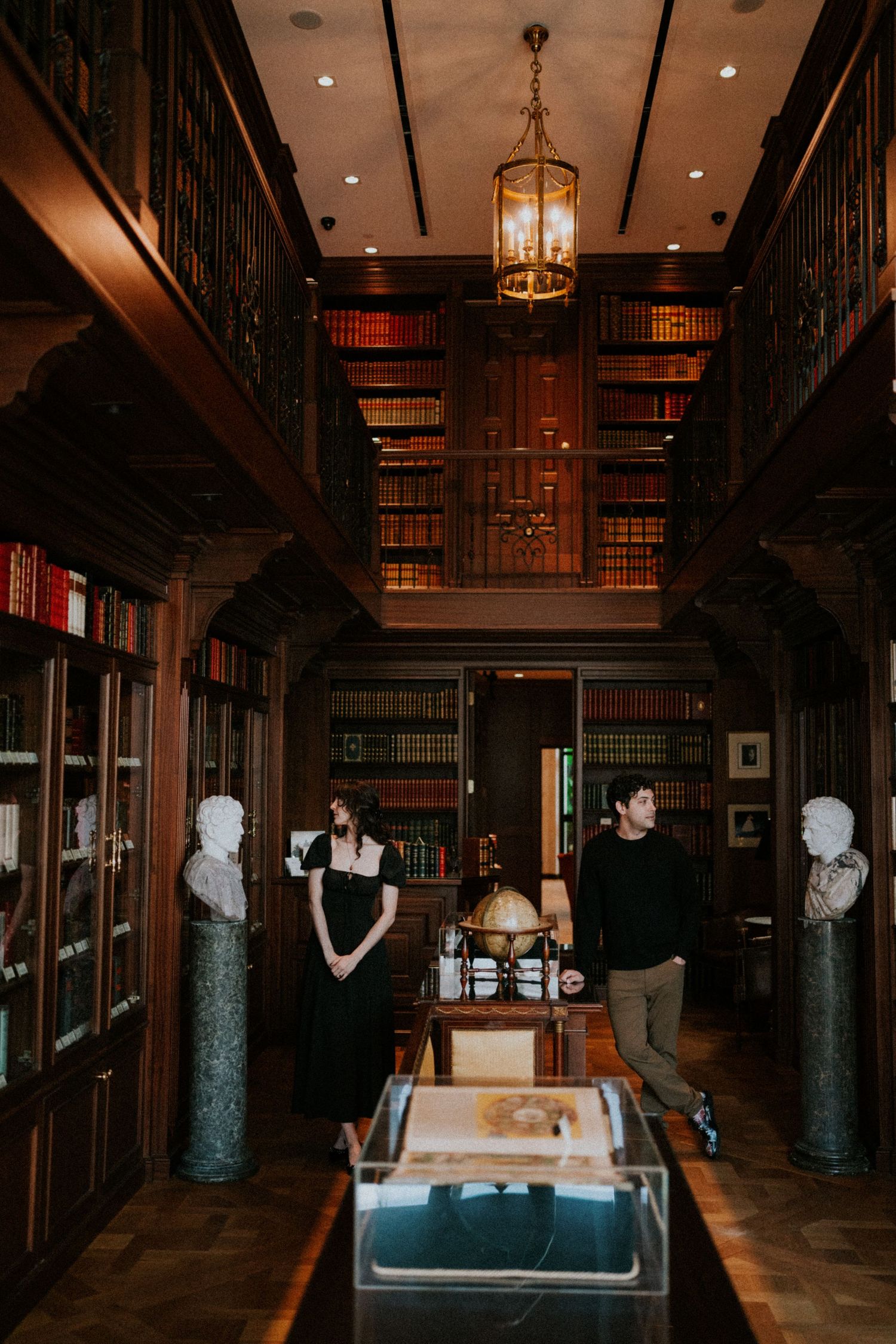 Two people stand in an elegant wood-paneled library with floor-to-ceiling bookshelves, marble busts, and a vintage chandelier.