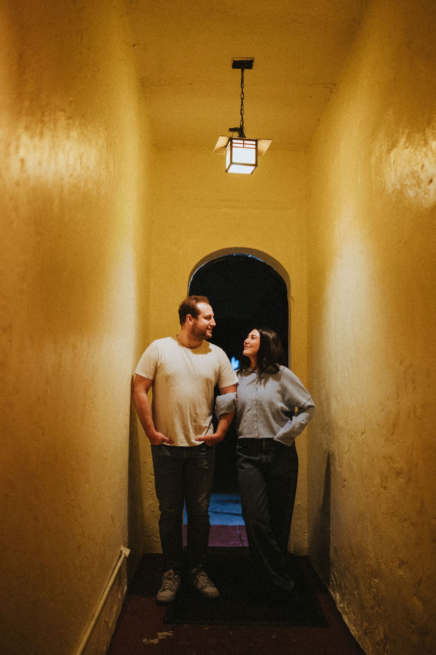 Couple smiling at each other in a narrow warmly lit hallway with yellow walls and an arched doorway.