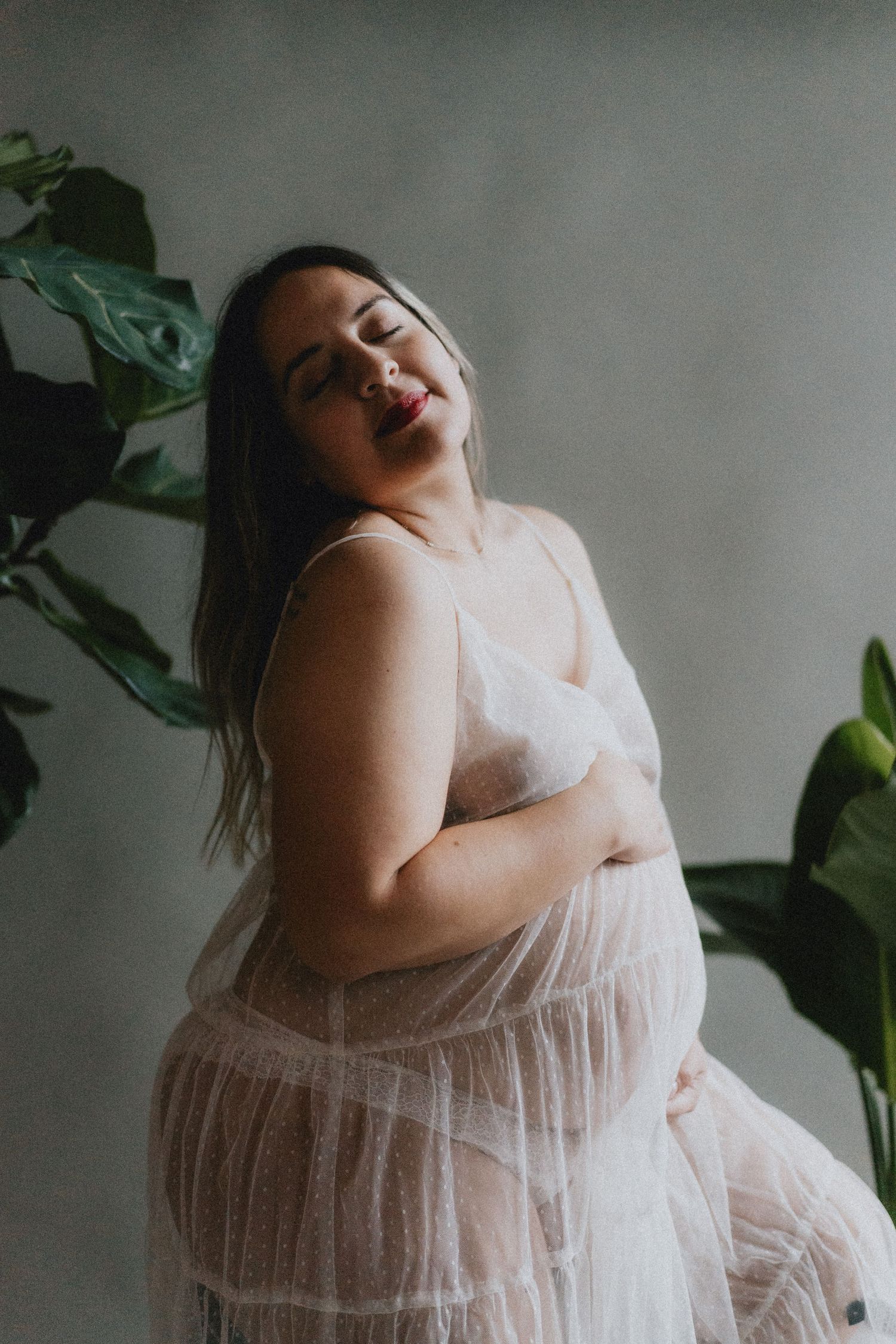 A person in a flowy white lace maternity dress poses against a grey wall with green plants in the background.